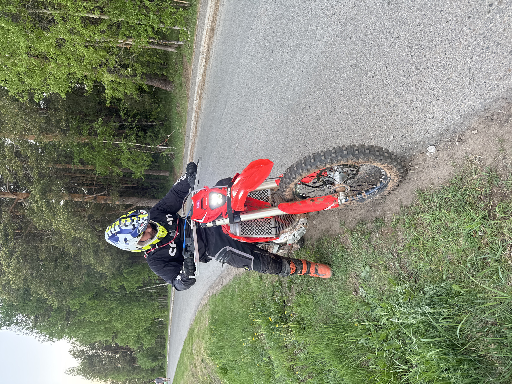 The image shows a person on a red dirt bike parked on the side of a road. The person is wearing a helmet, riding gear, and orange boots. The road is paved, and there is a grassy area next to the road. There are trees in the background. Given the location of Прикамский, Russia, the surrounding area would likely feature mixed forests.