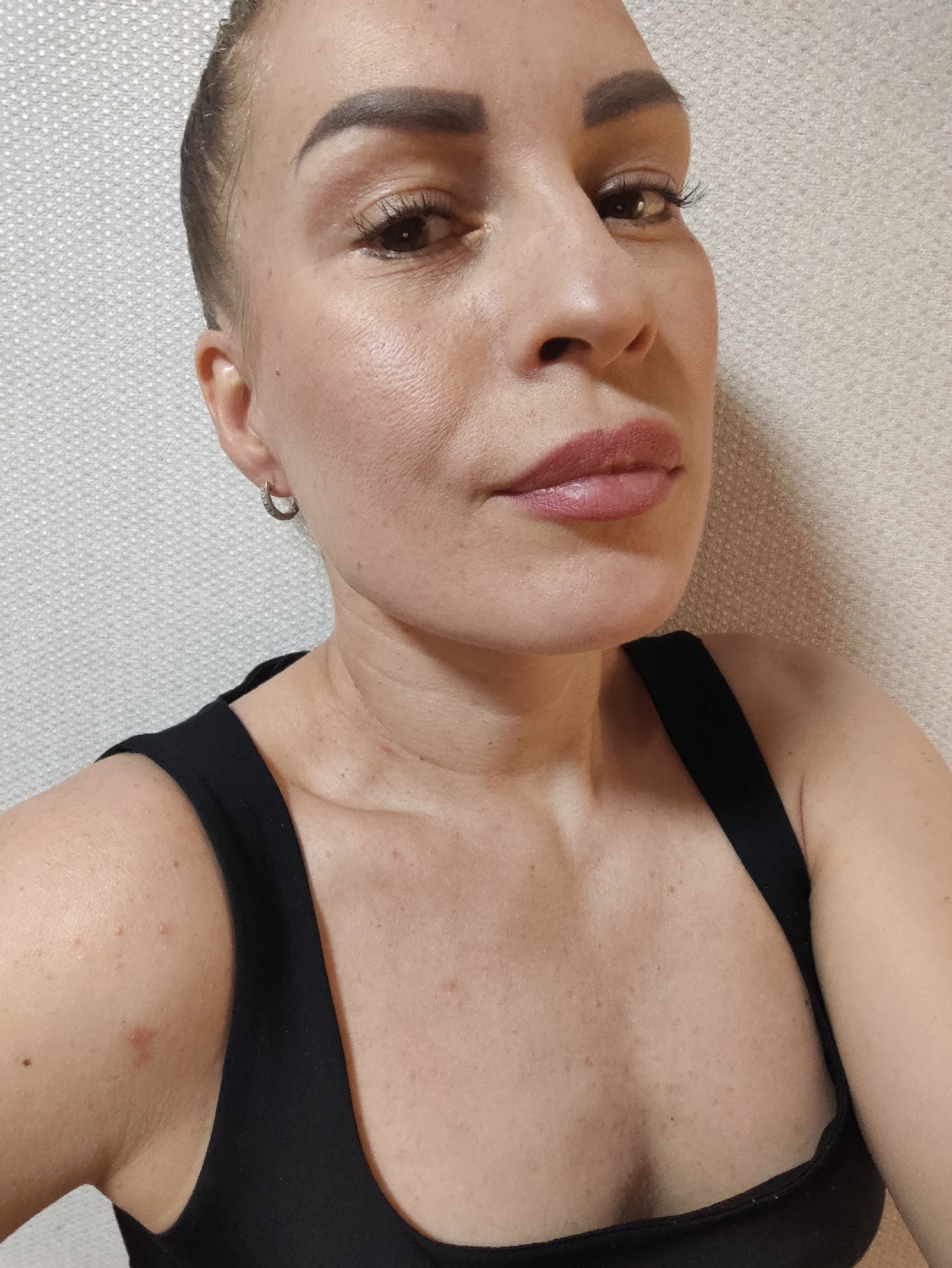 A close-up, medium shot focuses on the face and upper chest of a woman with light skin and dark hair pulled back. She is looking upwards and slightly to the right, with a neutral expression. She wears a black tank top. The background is a textured, light-colored wall.
Notable details include:
* **Eyes:** Her eyes are open, with dark irises and visible eyelashes. She appears to be wearing eye makeup, including mascara and eyeliner.
* **Eyebrows:** Her eyebrows are dark and well-defined, with a slight arch.
* **Lips:** Her lips are full and appear to be wearing a matte lipstick in a muted pink or mauve shade.
* **Skin:** Her skin shows some texture and a few small blemishes, particularly on her chest and shoulder.
* **Jewelry:** She is wearing a small, silver hoop earring in her left ear.
The lighting is soft and appears to be from an artificial source, possibly indoors given the plain, textured background and lack of distinct shadows associated with natural light. There is no indication of the time of day or weather conditions. The setting appears to be an indoor environment, likely a home or studio, based on the simple background and close-up framing. No text is visible in the image. The overall mood is neutral and direct.