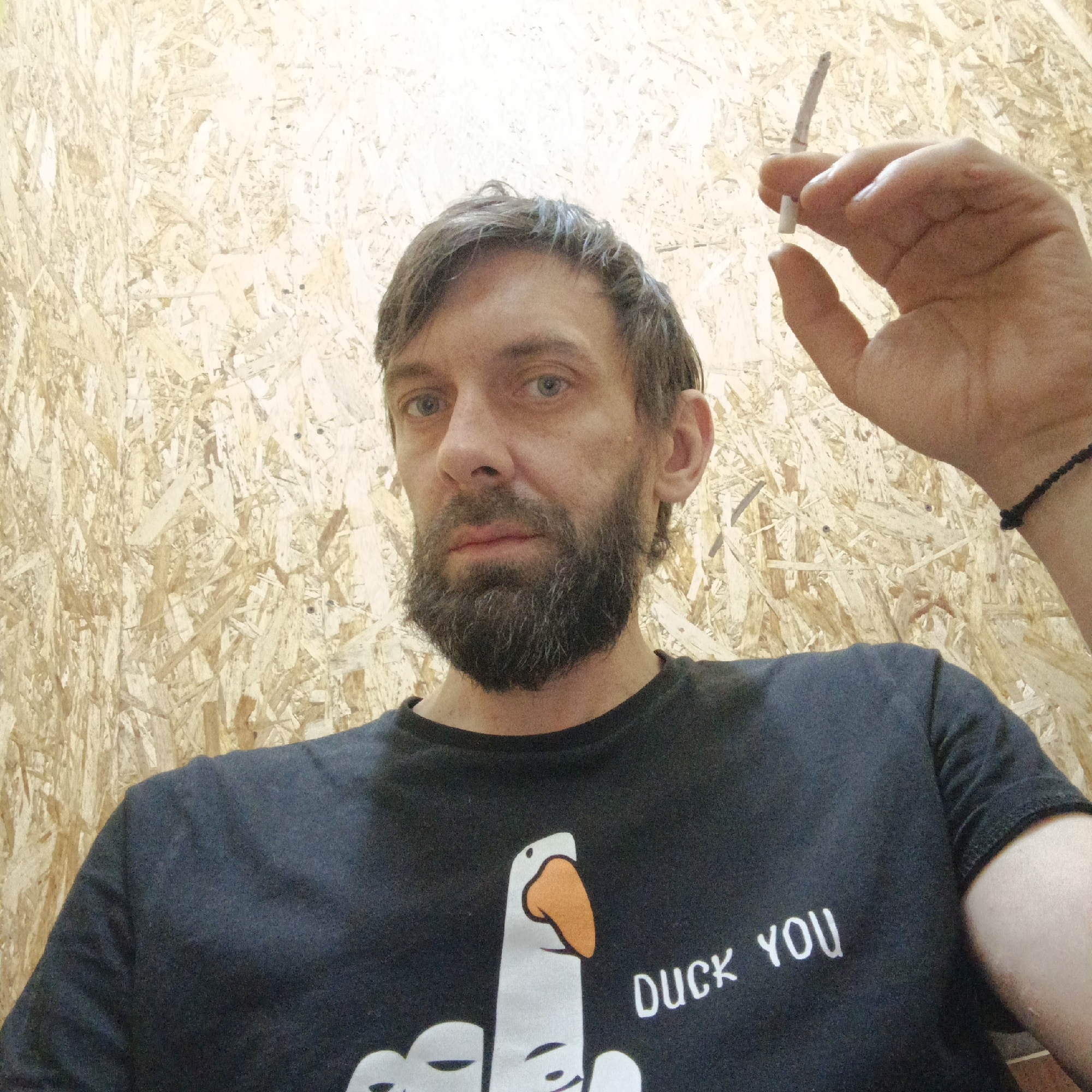 A man with a beard and dark hair is holding a lit cigarette in his right hand. He's wearing a black t-shirt with a cartoon goose giving the middle finger, and the words "Duck You" printed next to it. The background is a wall made of oriented strand board (OSB). There is no verifiable information to indicate that the image was taken in the city of Chaikovsky, Russia.