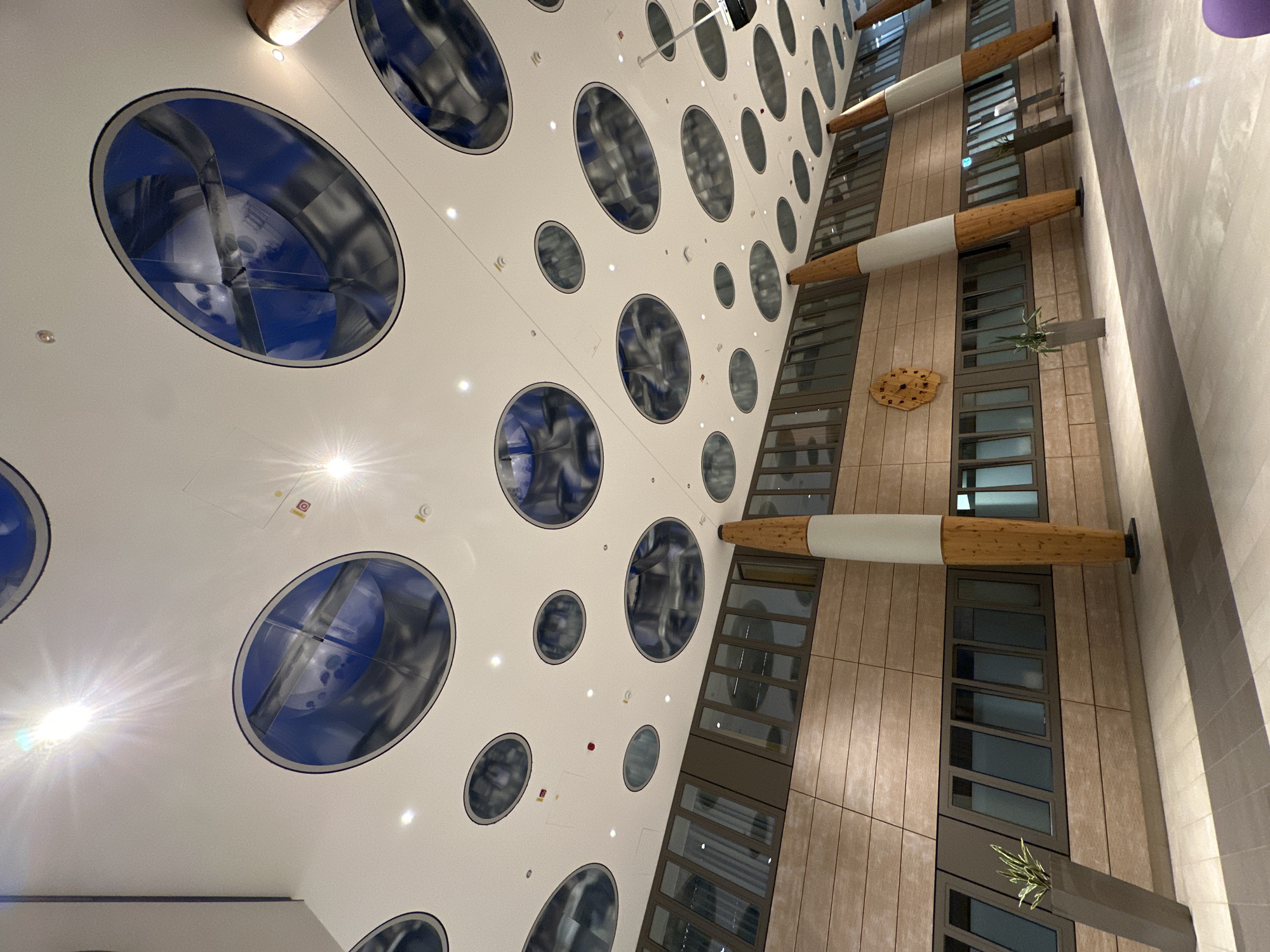 The image depicts the interior of a modern building, likely a public space or office, characterized by its striking ceiling design and warm-toned wooden paneling on the walls. The ceiling is a prominent feature, with numerous large, oval-shaped illuminated panels that are a deep blue color and appear to have a reflective or metallic surface, creating an abstract, almost celestial effect. Small, white recessed lights are interspersed between these larger panels. The walls are covered in horizontal wooden planks, giving a natural and sophisticated feel to the space. Two large, conical pillars, clad in wood with white accents, rise from the floor to the ceiling, providing structural support and adding a unique visual element. In the mid-ground, a series of large windows with dark frames run along the wall, reflecting the interior lights. To the left, a tall, dark gray planter contains a single green plant. A circular wooden clock with an intricate design is mounted on the wooden wall. The floor is a light-colored tile, with a darker gray pathway leading through the center of the space. The overall impression is one of contemporary architecture with an emphasis on natural materials and artistic design. This scene is located in the city of Garmisch-Partenkirchen, Germany.