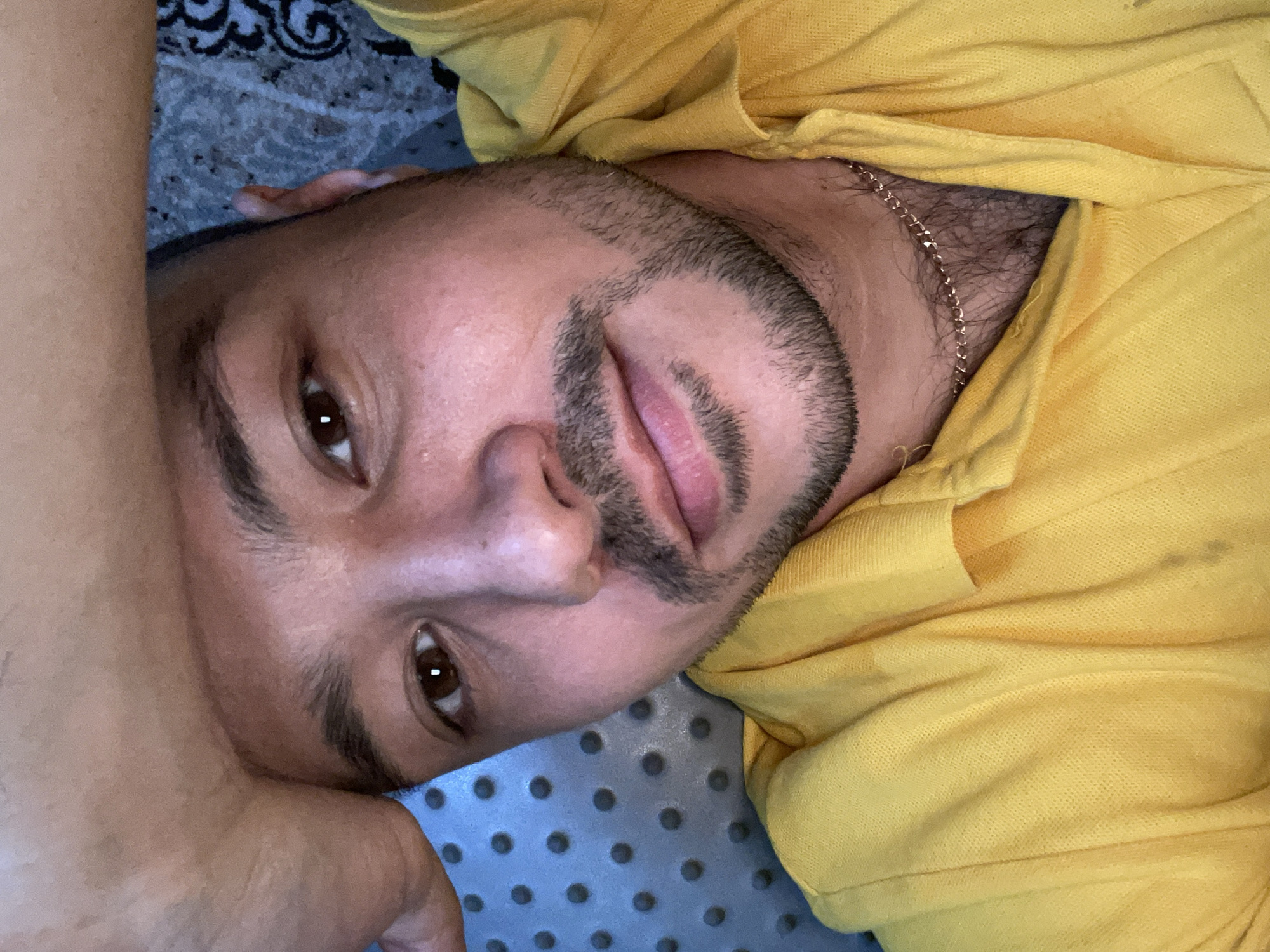 A close-up, inverted, selfie of a man lying on his back. He has brown eyes, dark hair, a short beard, and is wearing a yellow collared shirt with a gold chain visible at the neck. His left arm is bent, and his hand is near his head. He is lying on a patterned surface, which appears to be a mat or bedding. The background is out of focus. There is no evidence to suggest a specific location, including Zav'yalovo, Russia.