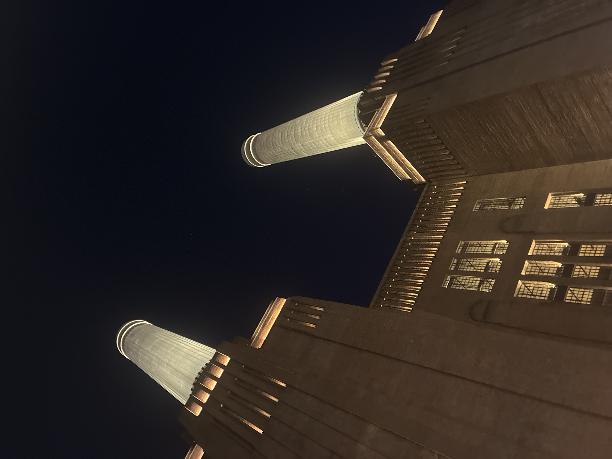 The image captures a low-angle view of the Battersea Power Station in London, United Kingdom, at night. Two of the power station's iconic chimneys are visible, illuminated against the dark sky. A section of the building's facade is also visible, showing rows of windows and architectural details lit by artificial light. The sky is a dark blue.