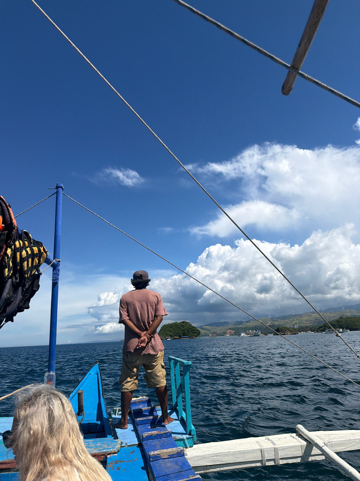 In Nabas, Philippines, a man wearing a maroon t-shirt and khaki shorts stands on the deck of a blue boat, facing away from the camera with his hands clasped behind his back. He is looking out at the choppy, deep blue ocean under a bright sky filled with white, fluffy clouds. To his left, a blue pole supports several life vests. In the distance, a chain of islands, some with lush green vegetation and wind turbines, lines the horizon. The foreground of the boat is visible, featuring blue wooden planks and a white outrigger. A woman with blonde hair is partially visible in the lower left corner of the frame, also on the boat. The scene captures a moment of quiet observation during a boat trip.