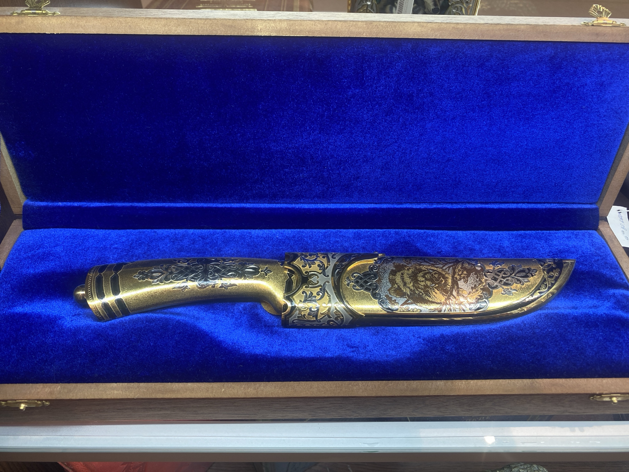 A highly ornate, gold-plated knife is displayed in a wooden box lined with blue velvet. The knife features intricate dark scrollwork on the handle and a detailed engraving of a tiger's head on the blade. The box is open, revealing the blue velvet interior and the knife resting within. The background shows the inside of the box, with hints of the wooden exterior and hinges. The overall impression is one of luxury and craftsmanship. There is no visible indication of Mikhalkovo, Russia in the image.