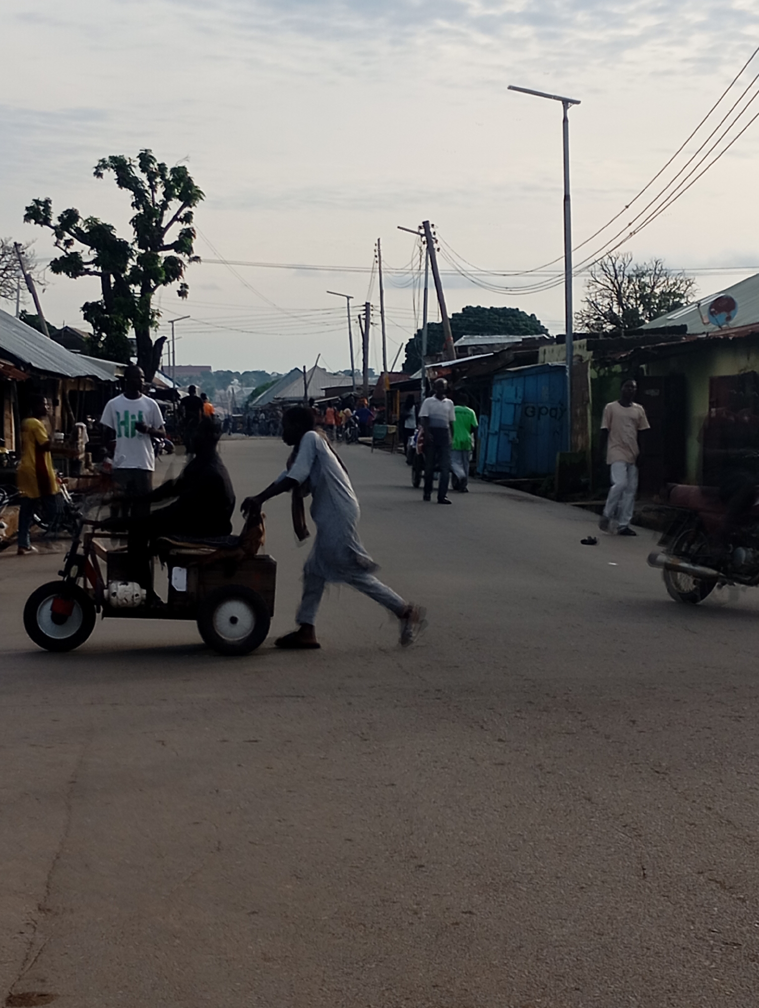 The image depicts a street scene in Shendam, Nigeria. In the foreground, a three-wheeled cart is being pushed by a person wearing a light-colored, long-sleeved garment and pants. Another person, wearing a white t-shirt with green lettering, is seated on the cart. Several other individuals are visible in the background, walking along the street. The environment appears to be an urban or semi-urban area with buildings lining the street, some with corrugated metal roofs. Power lines and streetlights are also present. The sky is overcast. The overall impression is a snapshot of daily life in a Nigerian town.