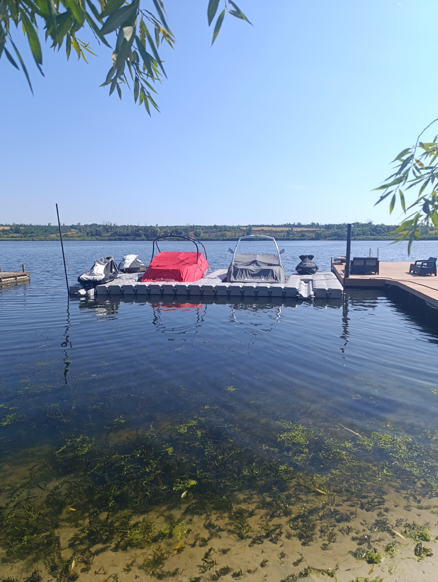 The image depicts a tranquil scene at the Bulatselivka reservoir in Bulatselivka, Ukraine. The reservoir, characterized by its clear, blue water, is dotted with aquatic vegetation along the sandy shoreline. A wooden dock extends into the water, serving as a mooring point for several watercraft. Among the vessels are two jet skis covered with protective tarpaulins and a small boat with a red canopy, also covered. A taller, grey boat with a metal frame is partially visible on the right side of the floating dock. The atmosphere is serene, with the calm water reflecting the clear blue sky and the surrounding lush green landscape. The sun casts a gentle light on the scene, highlighting the subtle ripples on the water's surface. The presence of vegetation and the calm waters suggest a peaceful, recreational setting.