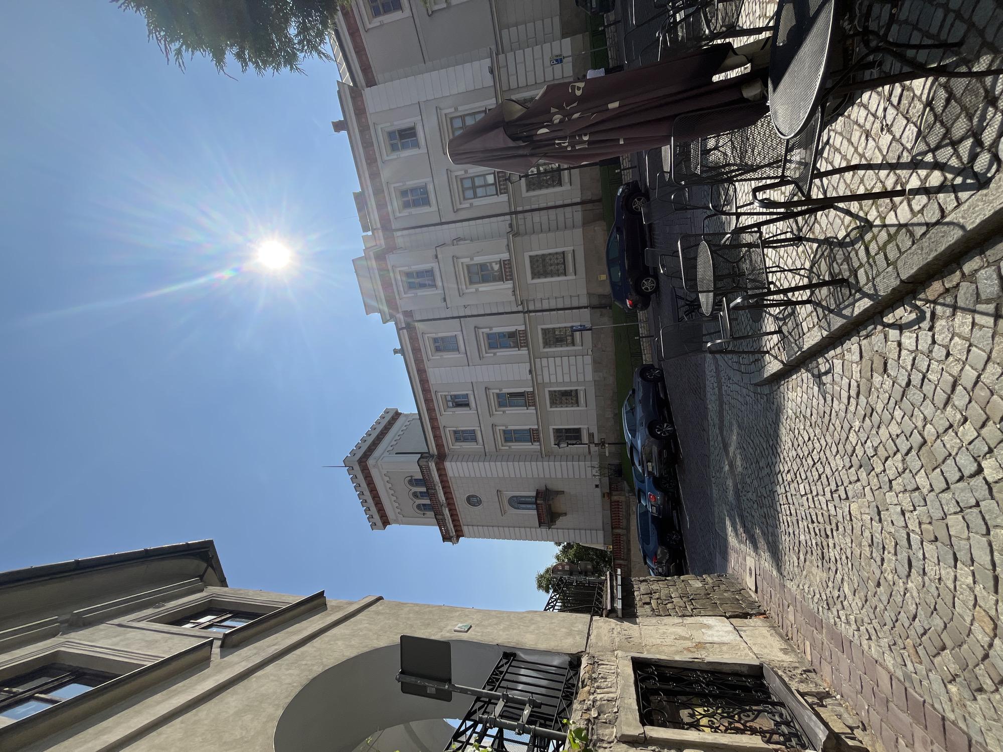 The image shows a street scene in a European city under a clear blue sky with a bright sun. In the foreground, a cobblestone street leads towards a large, ornate building with a tower. Several cars are parked along the side of the street. Outdoor cafe tables and chairs are set up on the sidewalk, with a large, closed umbrella dominating the right side of the frame. The building features classical architectural elements, including arched windows and decorative cornices. The overall impression is one of a sunny day in a historic urban environment.