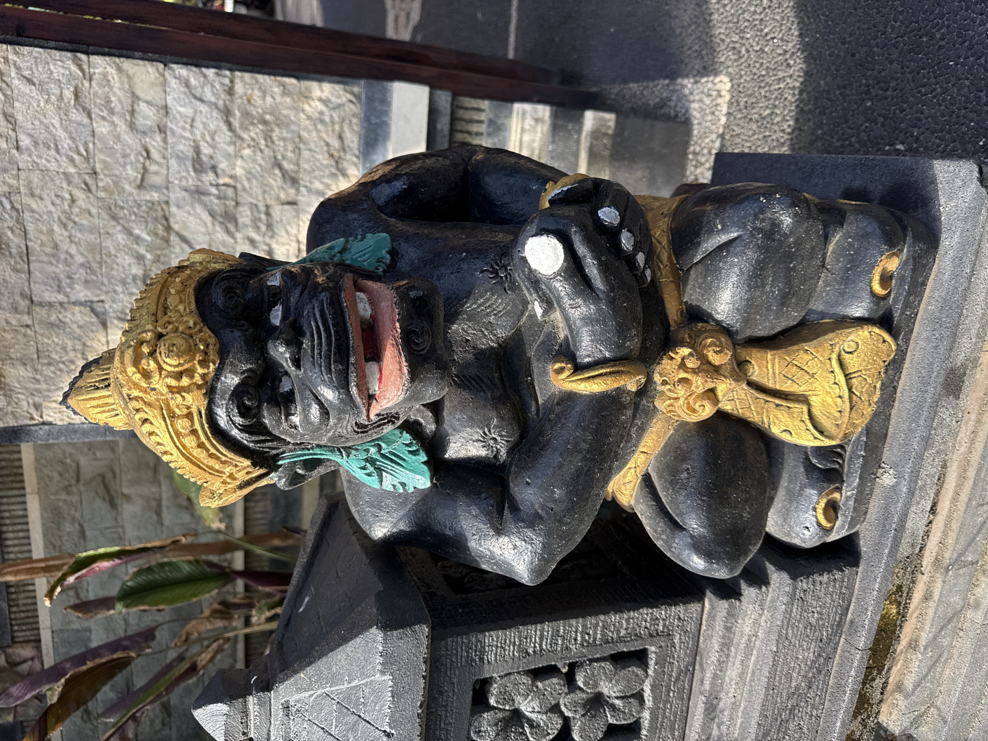 The image shows a statue in Kuta, Indonesia. The statue is dark gray with gold accents and features a figure with an open mouth, green accents around the neck, and a golden headpiece. The statue is placed on a dark gray, ornate pedestal. The background includes a stone wall, steps, and some foliage. The lighting suggests it is daytime with strong sunlight.