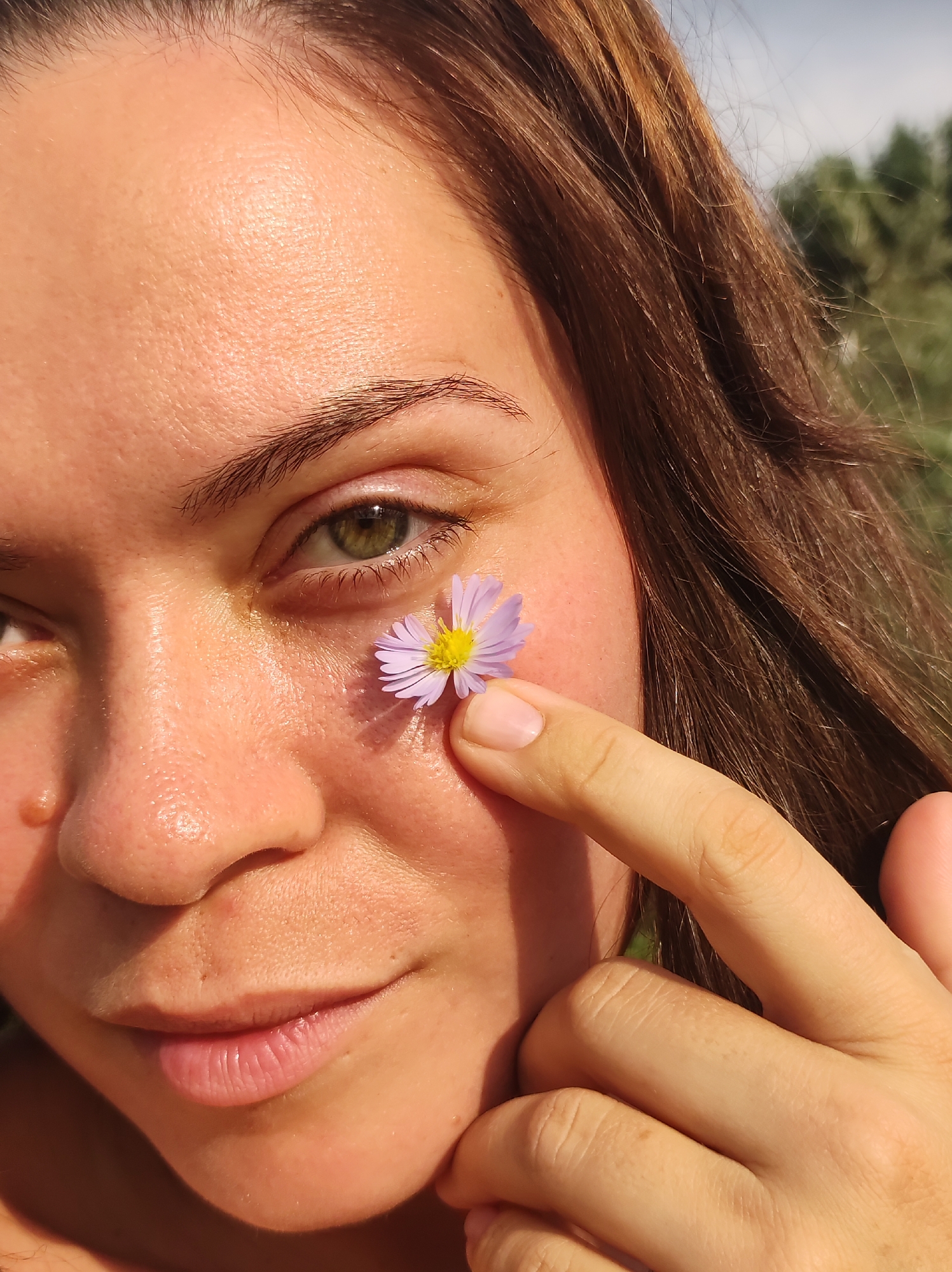 A close-up, outdoor, daylight photograph shows the right side of a young Caucasian woman's face. Her brown hair frames her face, and her green eyes are visible. She is holding a small, light purple flower with a yellow center against her cheek with her right index finger. The background is blurred, showing greenery and a bright sky, suggesting an outdoor setting possibly in a park or natural area. There are no visible landmarks that would indicate a specific city or country.