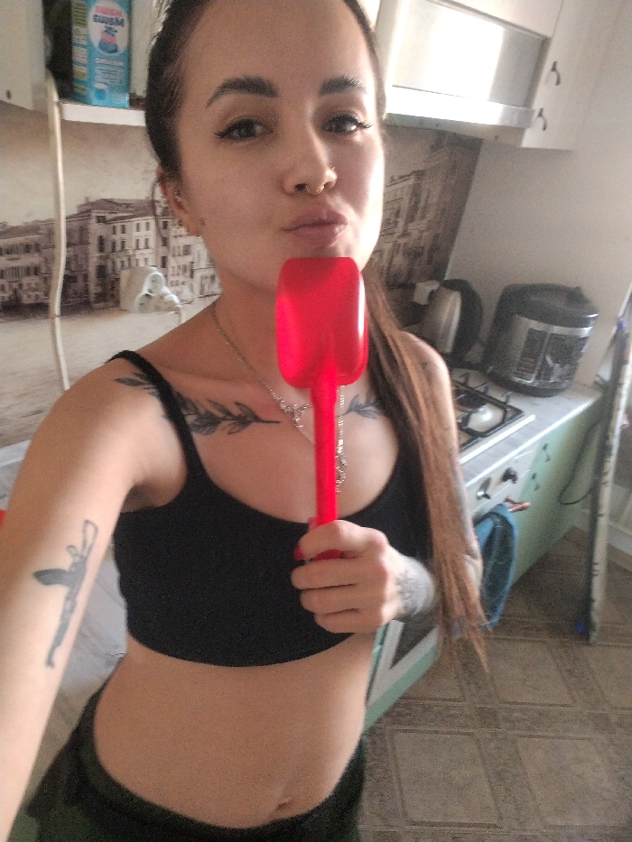 An image taken in a kitchen within the city of Noviy, Russia, depicts a female individual in a direct frontal pose, appearing to take a selfie. She has dark hair, a nose piercing, and multiple tattoos, including a leafy design on her upper chest and a stylized figure on her left bicep. She wears a black crop top, exposing her midriff, and dark lower garments, along with a silver-colored necklace. Her lips are pursed, and she holds a red, translucent, scoop-shaped utensil vertically in front of her mouth with her right hand.
The background reveals a domestic kitchen environment. To the subject's left, a wall features wallpaper depicting a sepia-toned cityscape, below which a white shelf holds a light blue and white package with Cyrillic text. To the subject's right, a countertop is visible with a two-burner gas stovetop, a black electric kettle, and a silver and black multi-cooker. Above the stovetop is a silver-colored range hood. Lower cabinets are light green, and a blue cloth hangs from one. The floor is tiled with a square, light-toned pattern, and a tall, white object, possibly a cleaning tool, stands upright on the far right.