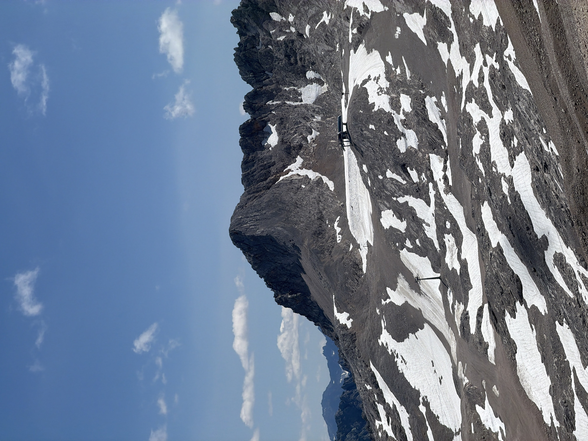 The image depicts a rugged mountain landscape with patches of snow on its slopes under a clear blue sky dotted with white clouds. In the foreground and mid-ground, rocky terrain is interspersed with snow, creating a stark contrast between dark grey rock and white snow. Several ski lift towers and cables are visible, indicating the presence of a ski resort or mountain transportation system. A building with a flat roof, likely a station for the ski lift, is situated on a higher plateau. In the background, more distant mountain ranges are visible, some with snow-capped peaks. The overall scene is one of a high-altitude alpine environment, likely during warmer months when snow is not widespread but still present at higher elevations. This scene is consistent with the Zugspitze mountain area near Garmisch-Partenkirchen, Germany, known for its glaciers, ski resorts, and cable car systems. No people are visible in the image.