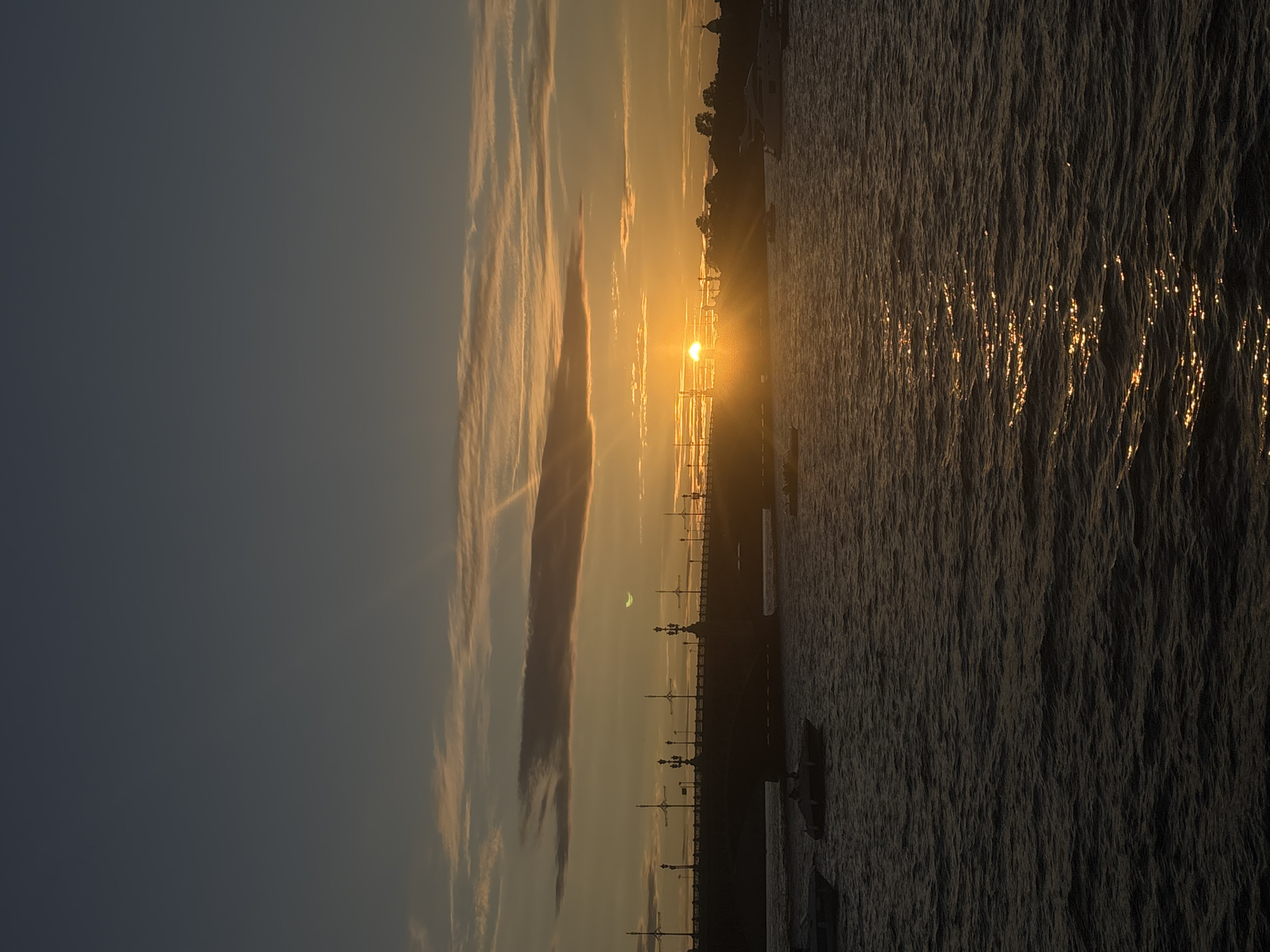 The image depicts a sunset over the Neva River in Saint Petersburg, Russia. The sun, a bright orb, is setting behind a bridge, casting a warm golden glow across the sky and reflecting on the rippling water. The bridge is silhouetted against the vibrant sky, its structure marked by ornate lampposts. Several boats are visible on the river, some docked and others navigating the water. The sky transitions from a deep blue at the top to shades of orange, yellow, and pink near the horizon, with wispy clouds catching the last rays of sunlight. The overall scene is serene and picturesque, capturing the beauty of a summer evening in Saint Petersburg.