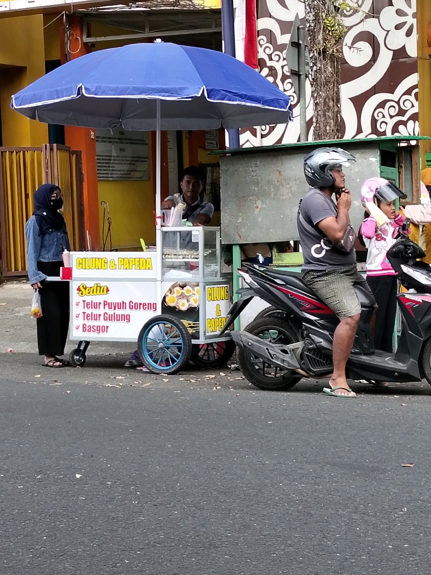 In Purworejo, Indonesia, a street scene unfolds with a food cart as the central focus. The cart, adorned with a blue umbrella, displays a sign that reads "CILUNG & PAPEDA" and advertises "Sedia: Telur Puyuh Goreng, Telur Gulung, Basgor" (Available: Fried Quail Eggs, Rolled Eggs, Basgor). A vendor, a man wearing a grey t-shirt and patterned shorts, is seated on a black motorcycle, adjusting his helmet. Beside him, a young girl in a pink jacket and helmet is also on the motorcycle. To the left of the food cart, a woman wearing a hijab, denim jacket, and face mask stands holding a plastic bag containing food. The motorcycle is parked on an asphalt road, with debris scattered on the ground. Behind the food cart, a yellow building with an orange fence is visible, and further back, a building with a brown and white patterned mural and green accents can be seen. The scene is captured in daylight.