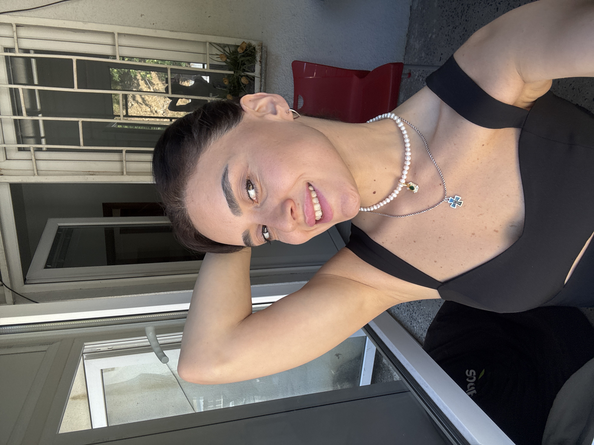 A woman with dark hair pulled back, wearing a black top and pearl necklace, smiles at the camera with her right arm raised behind her head. She is positioned in front of a large window with a metal grate on the exterior, suggesting an urban setting. The background includes a red chair, a window with a view of foliage, and a glimpse of a doorway. The lighting indicates it is daytime, likely outdoors or in a well-lit interior space near a window. The presence of a window with a grate and the architectural style of the building are consistent with urban environments such as Tbilisi, Georgia.