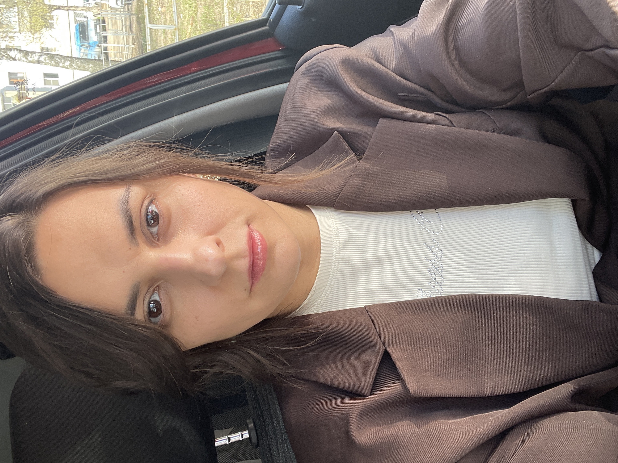 Here is a description of the image:
A woman is in a vehicle. She is wearing a brown jacket over a white top. Outside the car is a street view that includes a white building and a light-colored truck.
