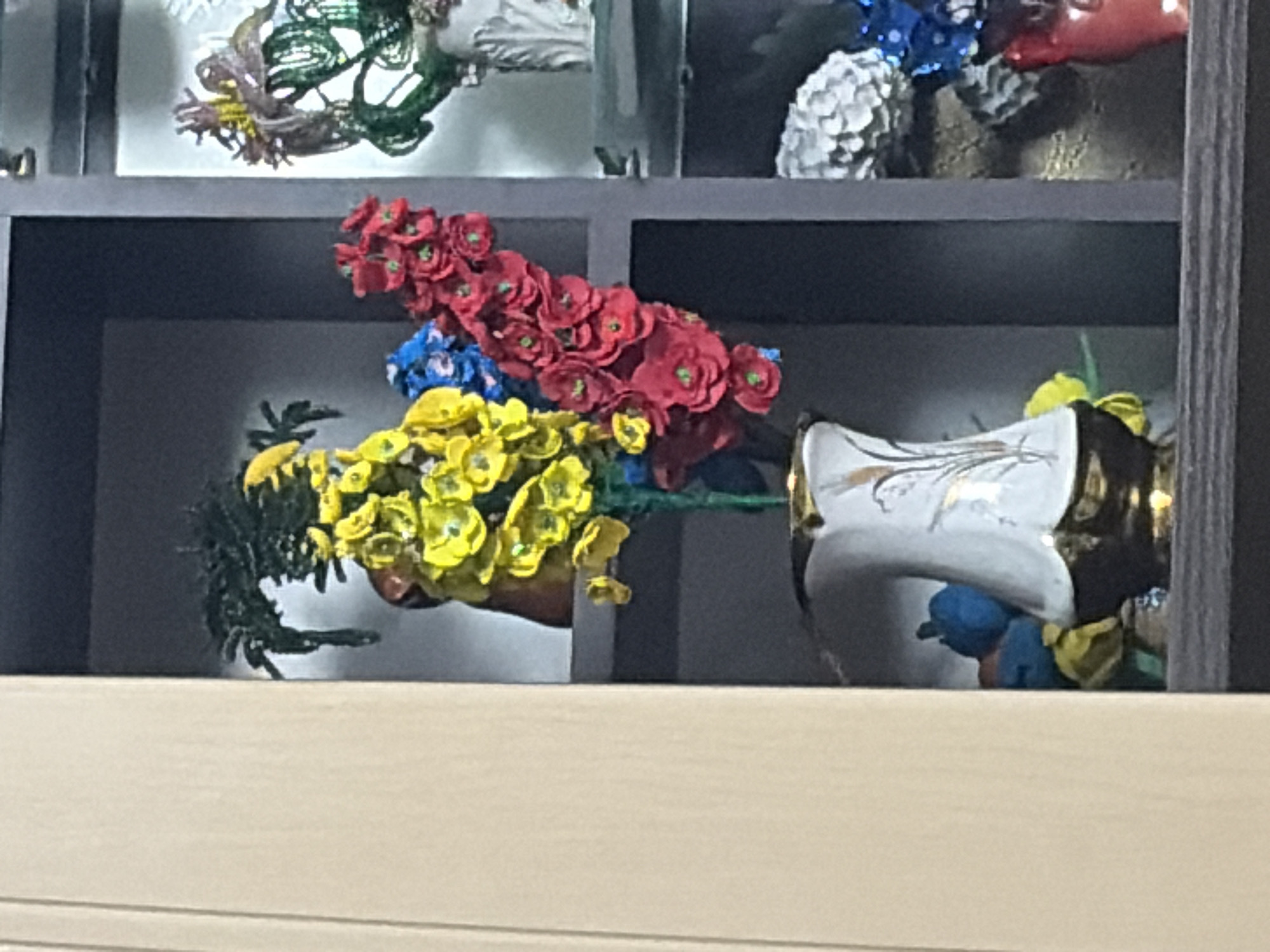 The image shows a decorative floral arrangement in a vase, displayed on a shelf in what appears to be an interior setting. The arrangement features clusters of bright yellow, blue, and red artificial flowers. The vase itself is white with gold accents and a metallic gold base. Behind the main floral arrangement, other decorative items are visible, including what looks like a beaded green floral display on a higher shelf and a white, textured floral arrangement and a red vase on a lower shelf. The background is a dark gray shelving unit, with a lighter colored wall or cabinet visible to the left. The lighting suggests an indoor environment, possibly a home or a shop display. There are no people or animals present. The location of Chaikovsky, Russia is not discernible from the visual information within the image.