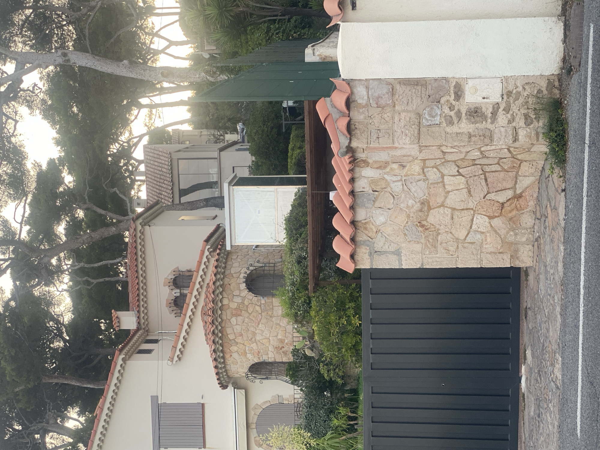 This is a photograph of a multi-story house in Antibes, France, with a dark gray metal gate in the foreground. The house features a red tiled roof and is constructed with a combination of cream-colored walls and a stone-faced turret. The turret has two arched windows with decorative iron grates. The surrounding vegetation is lush and green, with several tall pine trees visible behind the house. The sky is bright and appears to be late afternoon or early evening. A white-painted pillar is to the right of the stone wall in the foreground. There are no people visible in the image.