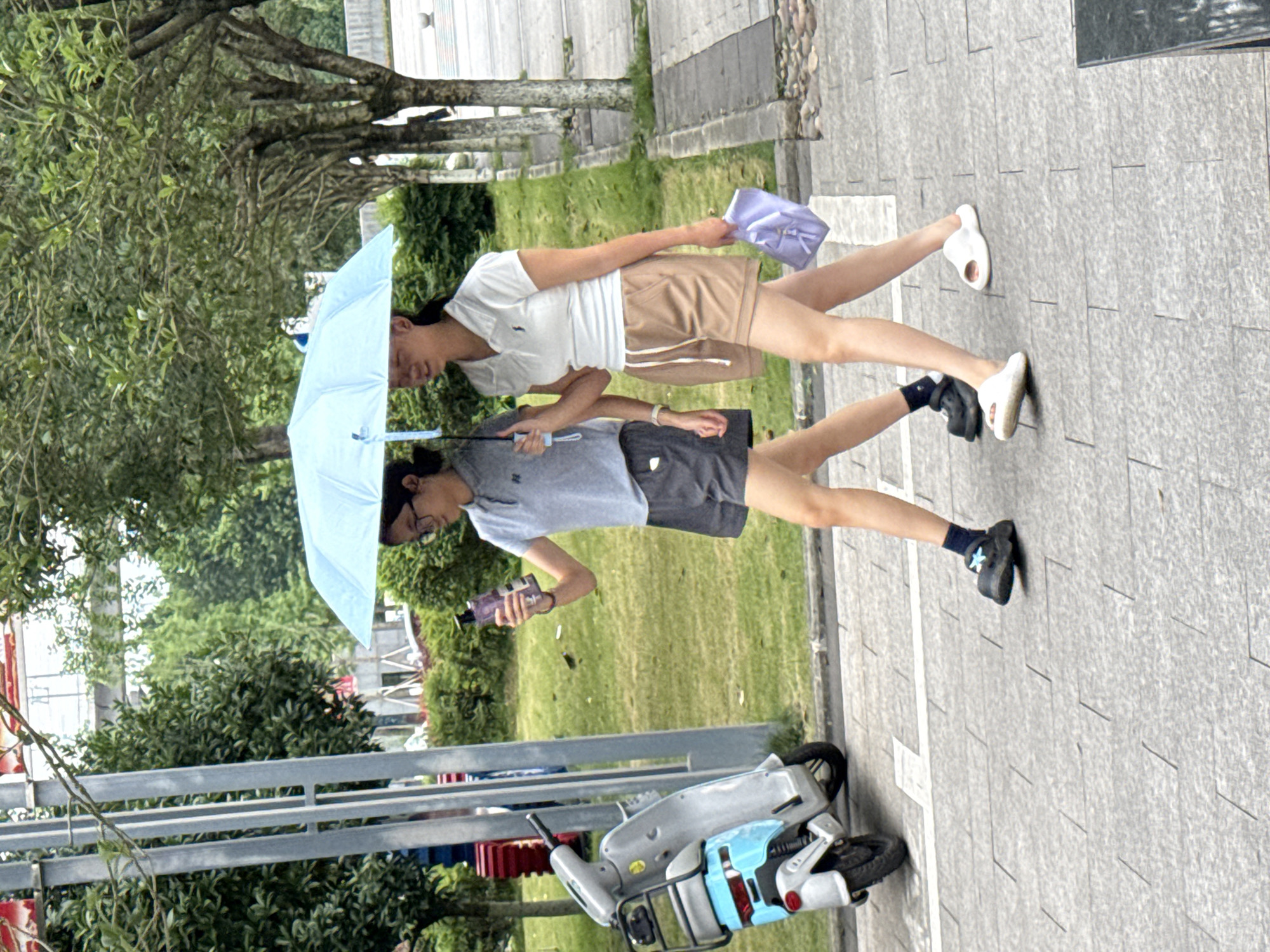 Two individuals walk side-by-side under a light blue umbrella on a paved path in what appears to be an urban park setting in Jiangjin, China. The person holding the umbrella is wearing glasses, a light grey t-shirt, dark shorts, and black sneakers with socks. They are carrying what looks like a bottle. The other person is wearing a white polo shirt, brown shorts, and white sandals, carrying a small purple bag. The path is bordered by grass and trees. An electric scooter is parked near the bottom left of the frame. The pavement has linear markings.