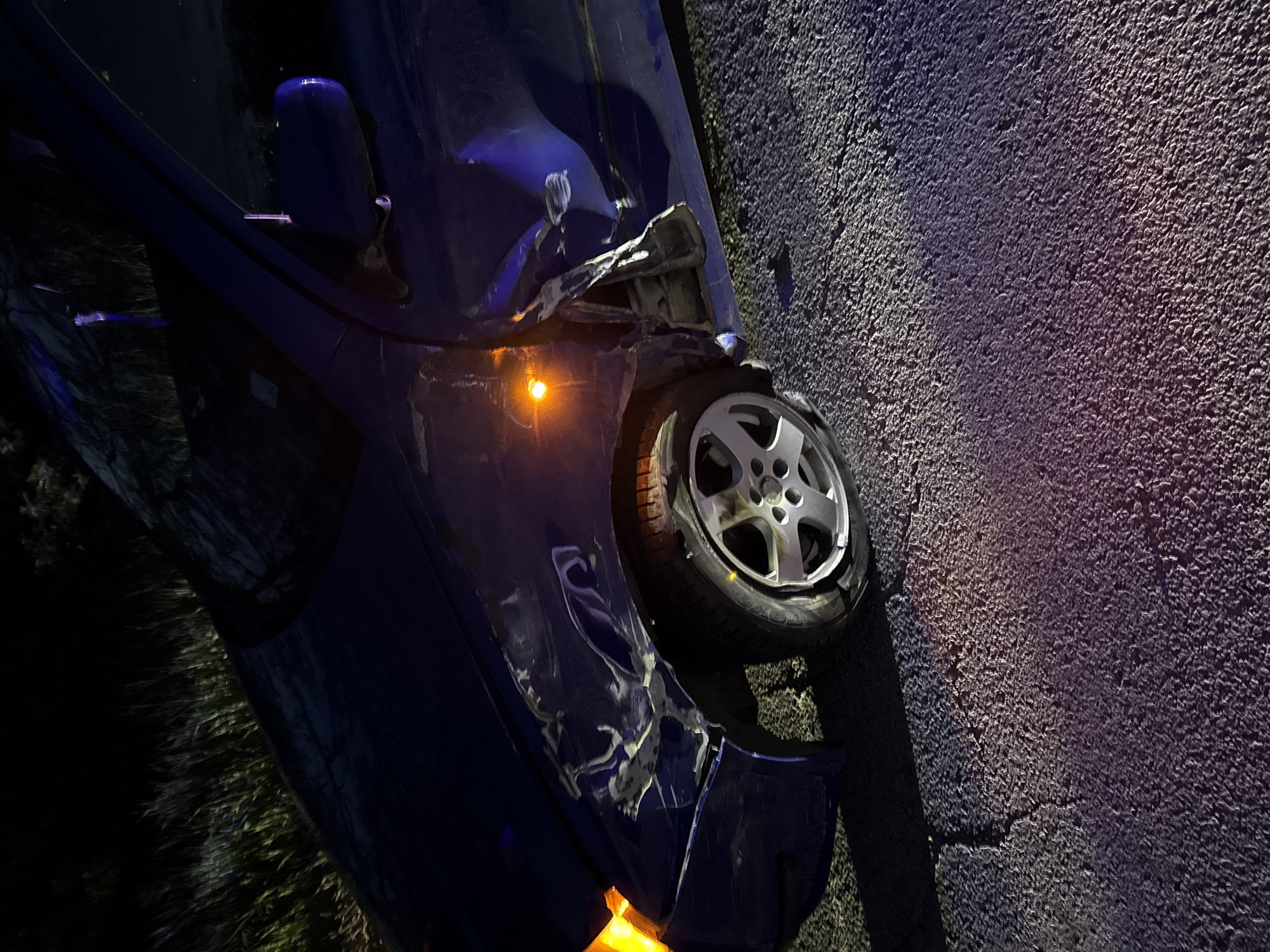 In the image, a damaged blue car is seen on what appears to be an asphalt surface. The front right side of the car shows significant damage, with the front right wheel appearing bent or misaligned. The turn signal is illuminated, casting an orange glow. The car's body has visible scratches and dents. The background and surroundings are dark, suggesting the incident occurred at night. There is some vegetation visible near the front of the car. While the prompt asked for the location in Opanets, Bulgaria, this is not discernible from the image. The scene appears to be a car accident.