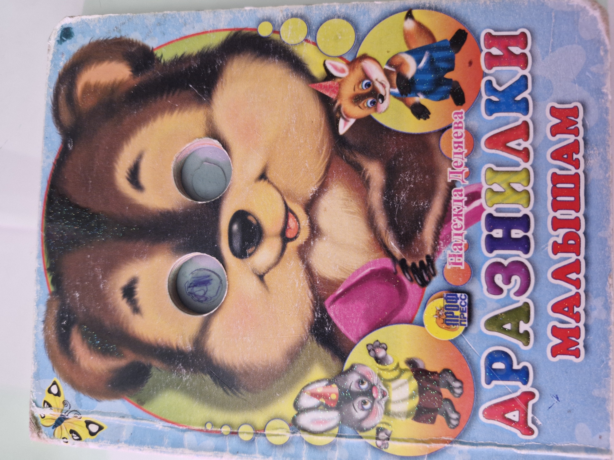 The image shows the cover of a children's book titled "ДРАЗНИЛКИ МАЛЫШАМ" (Teasers for Little Ones). The book features a cartoon bear with two circular cutouts for eyes, through which a child's drawing is visible. To the left of the bear is a cartoon rabbit wearing a party hat and a yellow shirt, and to the right is a cartoon fox in a blue jumpsuit and a party hat. The book cover also includes a small circular logo with Russian text and a butterfly illustration in the upper left corner. The overall condition of the book cover suggests it is old and has been used, with visible wear and tear, and some glitter embellishments. There is no discernible information within the image that would allow for a determination of its location in Kazan, Russia.