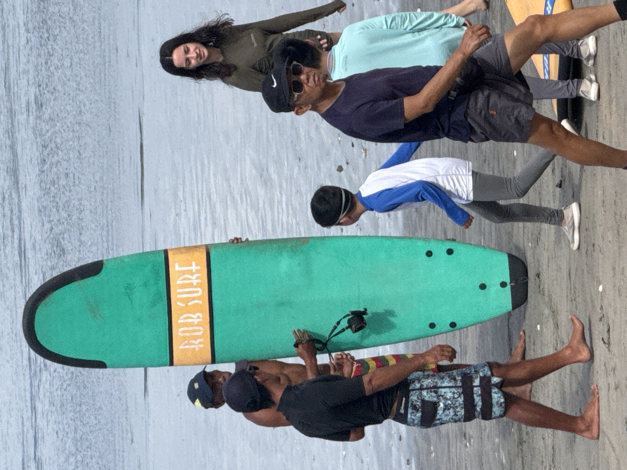 In Kuta, Indonesia, on a beach with calm water in the background, several people are gathered near a large, green surfboard. A man wearing a black shirt and patterned swim trunks walks in the foreground, facing left. Behind him, a man holding the surfboard also faces left, wearing a black cap and swim trunks. Another man, partially visible, stands next to the surfboard. In the mid-ground, a person wearing a blue and white long-sleeved shirt and grey leggings walks to the left. To the right, a woman with dark hair stands looking down, wearing a long-sleeved olive green top. Another person, wearing a dark blue shirt and grey shorts, stands facing the camera. Further to the right, a person in a light blue long-sleeved shirt is also visible. The sand is wet, indicating the tide is close.