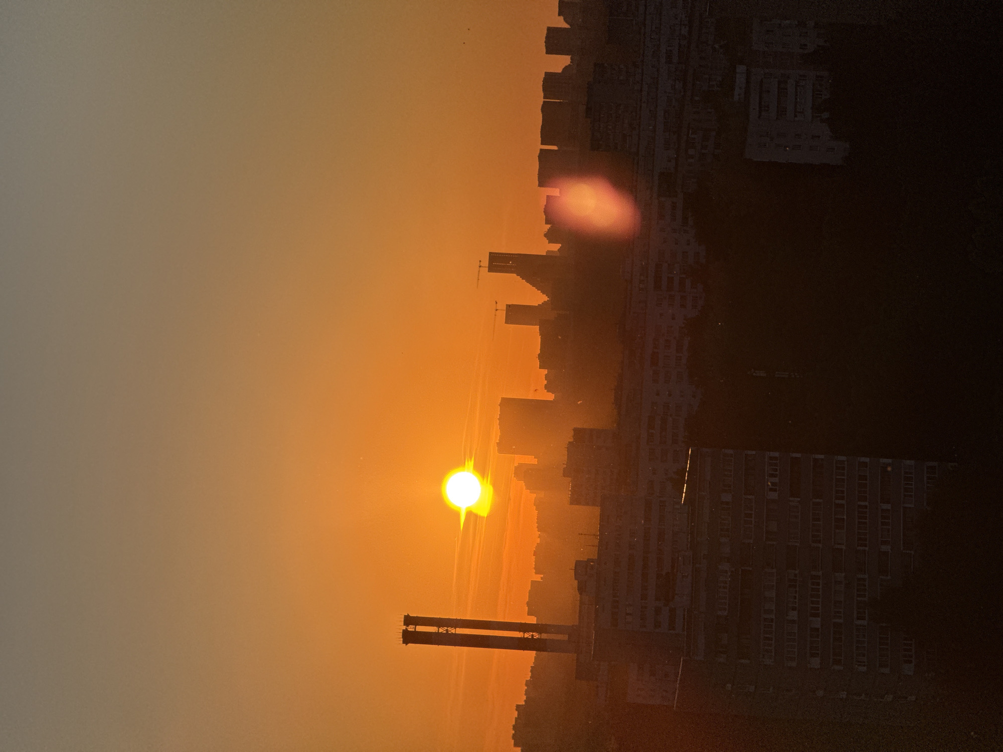 The image depicts an urban cityscape at either sunrise or sunset, characterized by strong backlighting. The sky transitions from a brilliant orange-yellow near the horizon, where the sun is visible as a bright, circular, white-yellow orb, to a muted greyish-brown in the upper portion. A prominent, reddish-orange lens flare is visible in the lower right quadrant.
The foreground and midground are dominated by the dark silhouettes of numerous multi-story residential buildings, consistent with typical Soviet-era panel construction found throughout Moscow. In the left midground, two tall, slender, light-colored smokestacks, characteristic of a thermal power plant, rise prominently against the illuminated sky.
In the central-right background, several modern high-rise buildings are silhouetted. One taller structure exhibits a distinct angled or stepped profile and features two visible tower cranes, indicating active construction. Another slightly shorter modern building is visible to its left, also potentially under construction with a smaller crane. No individual people or specific actions are discernible due to the distance and lighting conditions. The overall blend of Soviet-era residential architecture, modern skyscraper development (including ongoing construction), and thermal power plant infrastructure is highly characteristic of the urban environment within the city of Moscow, Russia.