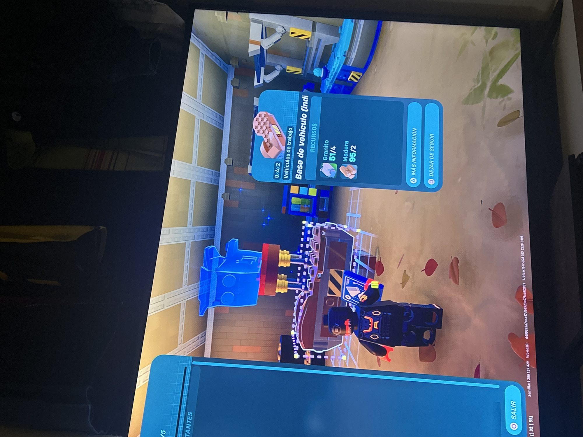 A photograph of a television screen shows a scene from a video game. The main focus is on a Lego-style character standing next to a construction made of Lego bricks. A blue, translucent blueprint appears to be overlaying part of the construction. A blue menu panel to the right of the Lego character displays information about a "Vehicle Base" (translated from the Spanish text). The panel lists the required resources as "Granite" with a 51/4 ratio and "Wood" with a 95/2 ratio. Two options are provided in the menu: "More Information" and "Stop Following." Another blue menu panel is visible at the bottom of the screen, with the word "SALIR" (Spanish for "EXIT"). The in-game environment consists of autumn leaves on a brown ground. There's also an in-game location displayed on the screen as "Ubicación:-328 763 2328 3195." The rest of the text on the screen appears to be game-related information.
