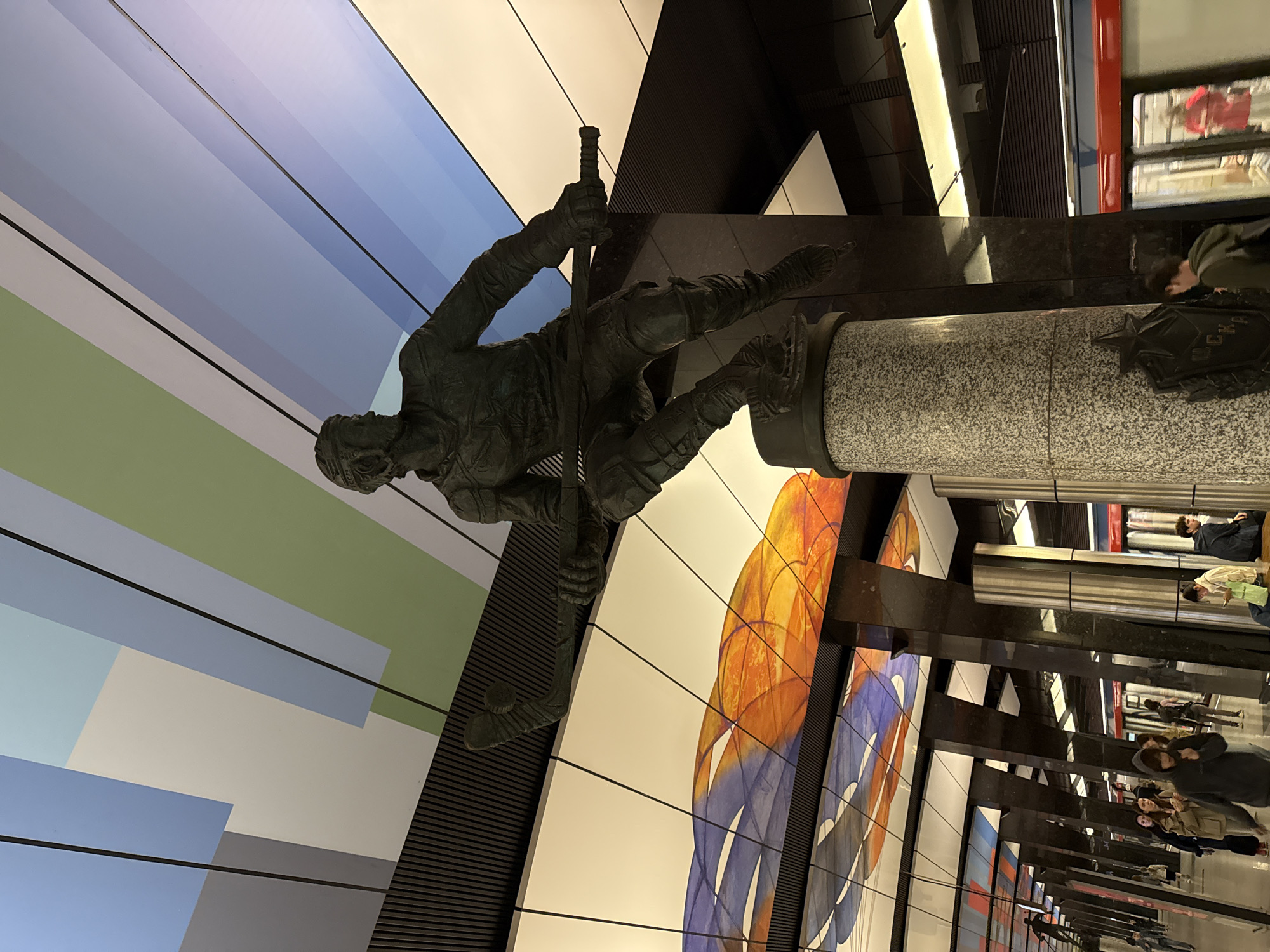 The image shows a sculpture of a soldier figure in a Moscow metro station. The sculpture is positioned on a cylindrical pedestal. The station has colorful abstract wall decorations, black structural supports, and a visible platform with people waiting for a train.