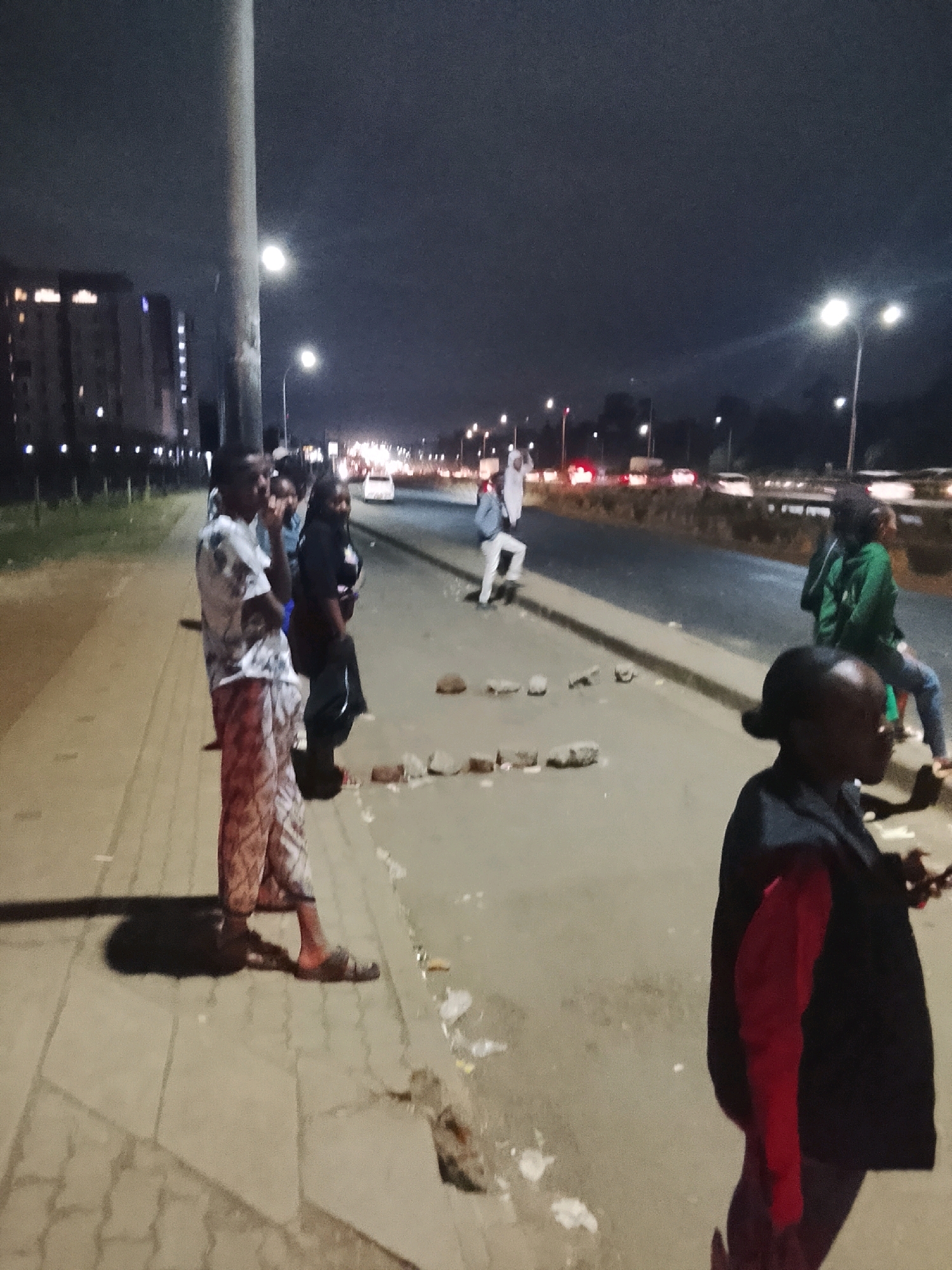 This is a medium-shot, night-time photograph taken on a sidewalk beside a busy road in Nairobi, Kenya. A group of people, appearing to be adults, are gathered on the sidewalk. One person in the foreground, on the right side of the frame, is wearing a black vest over a red top. To their left, a person in a patterned garment and sandals is standing. Further to the left, two individuals are standing close together, with one wearing a dark top and the other a lighter, patterned shirt. In the background, on the road, vehicles are driving by, their headlights and taillights creating streaks of light. Streetlights illuminate the scene, casting shadows and highlighting the dark, overcast sky. Several bricks are scattered on the ground on the sidewalk, near the edge of the road. The environment appears to be an urban setting with buildings visible in the distance. The overall mood of the image is somber and somewhat tense.