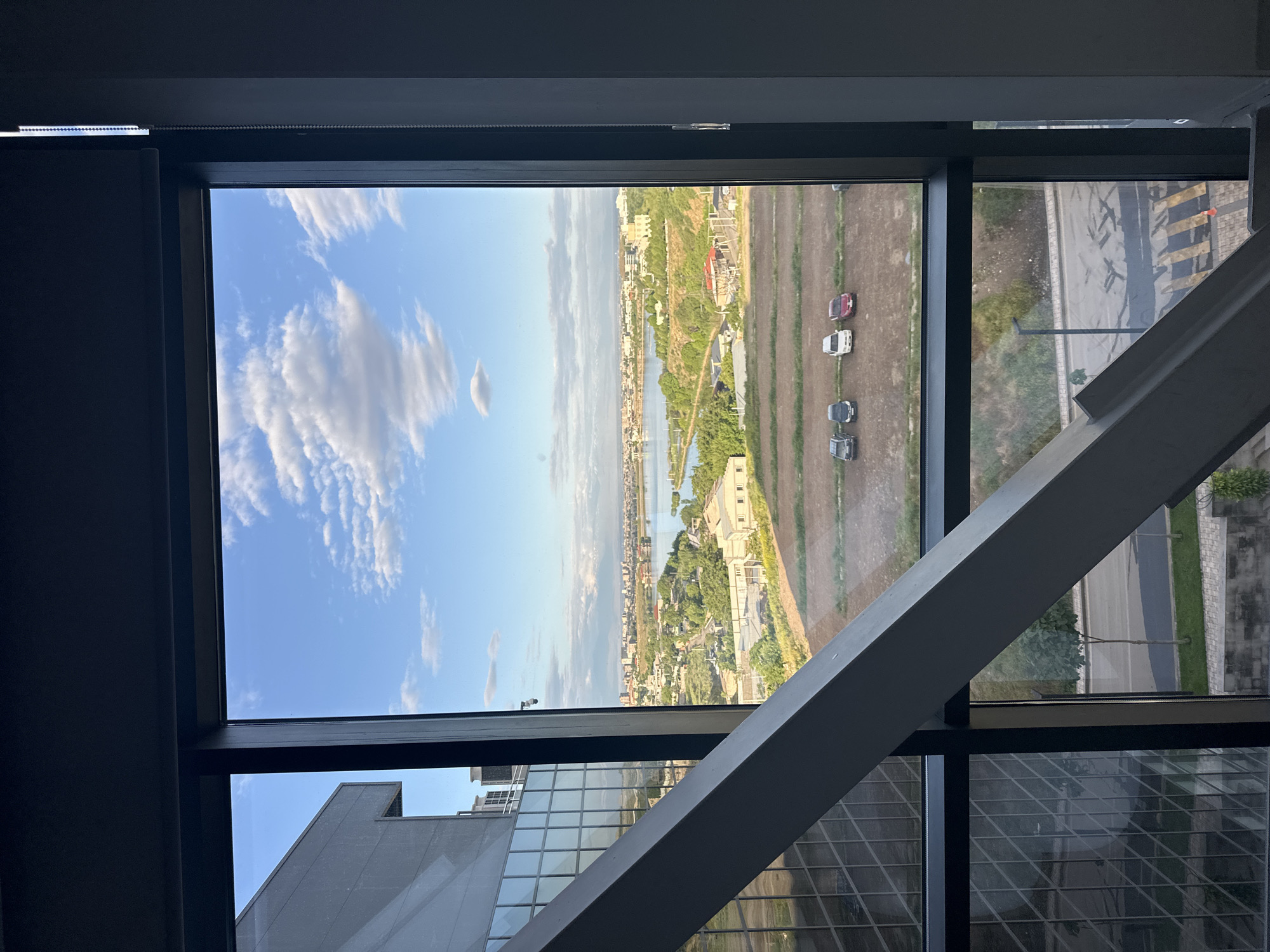 An interior view shows the cityscape of Yerevan, Armenia, through a large window. The window is framed in black and partially obstructed by a gray architectural beam angled across the lower right portion of the view. Outside, the sky is blue with scattered white clouds. In the distance, a body of water snakes through a landscape of buildings, greenery, and roads. Several cars are visible parked in a dirt area. The reflection of a nearby building can be seen in the window.