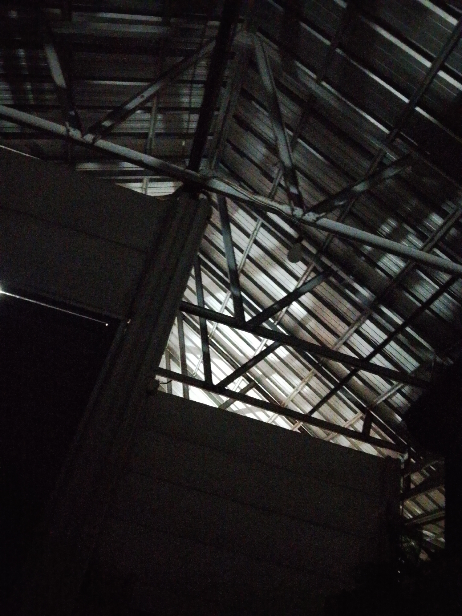 The image depicts the interior of a building, potentially a warehouse or large shed, in Bukit Ketok, Indonesia. The primary focus is the roof structure, composed of metal sheeting supported by a network of metal beams and trusses. The lighting is dim, casting shadows and obscuring some details. Part of an adjacent wall is also visible, appearing to be constructed from concrete or a similar material. There are no people or distinct objects other than the building's structure. The environment is dark and enclosed.