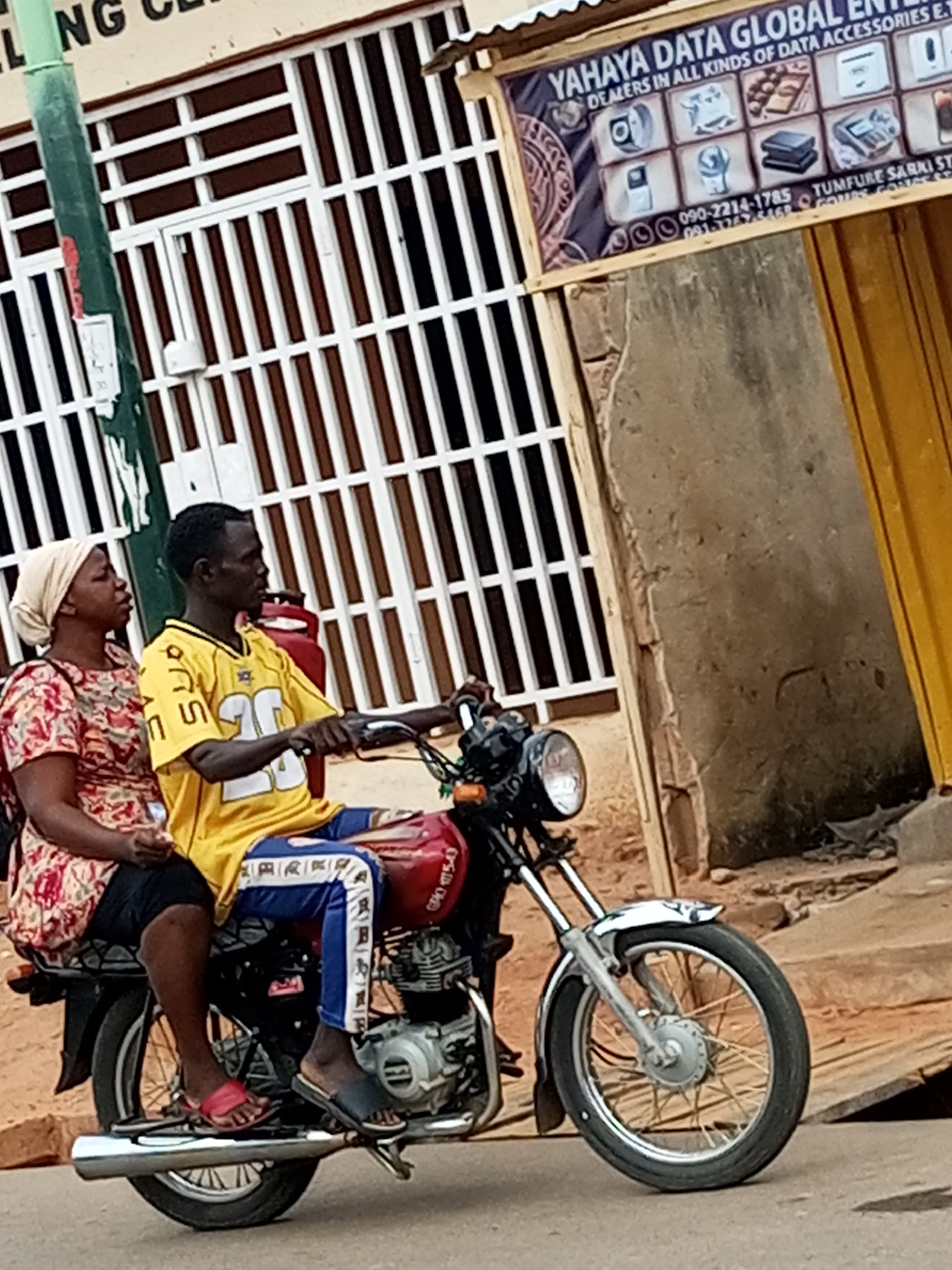 Two individuals, a man and a woman, are seated on a red motorcycle in the city of Tumfure, Nigeria. The man, positioned behind the handlebars, wears a yellow jersey with the number "26" and blue pants with white and blue stripes. The woman, seated in front of him, wears a patterned red and white shirt and a light-colored headscarf. The motorcycle's chrome exhaust pipe is visible, extending horizontally. In the background, a building features vertical white bars over a dark background and a sign for "YAHAYA DATA GLOBAL ENTERPRISES" which advertises data accessories and displays contact numbers. A green pole is also present to the left of the frame. The overall environment appears to be an urban street scene with a building and a road.