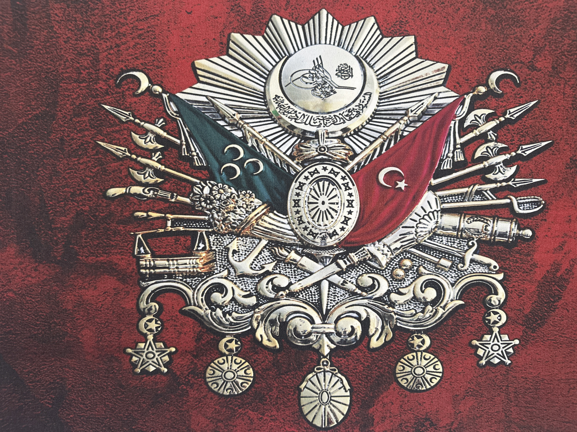 The image is a close-up, flat-lay photograph of a metallic emblem, likely a coat of arms or official seal, rendered in silver and gold tones against a textured, dark red background that resembles fabric. The emblem is highly detailed and features a central medallion surrounded by various symbols.
At the top center of the emblem is a circular disk with Arabic calligraphy, possibly a Tughra, above which rests a crescent moon. This central element is encased within a radiating sunburst pattern. Two flags are prominently displayed beneath the central medallion: one green with three crescent moons, and the other red with a white crescent moon and star.
Surrounding these elements are numerous weapons and symbols of power, including crossed swords, spears with ornate arrowheads, axes with curved blades, cannons, and an anchor. Decorative scrollwork and floral motifs form the lower part of the emblem, from which hang several ornate medallions and star-shaped ornaments.
The overall impression is one of historical significance and authority. The emblem appears to be a physical object, possibly a wall hanging or a decorative plaque.
The image does not contain any information about the city of Masazir, Azerbaijan, nor does it depict any people, actions, or interactions. It is solely a representation of the metallic emblem.