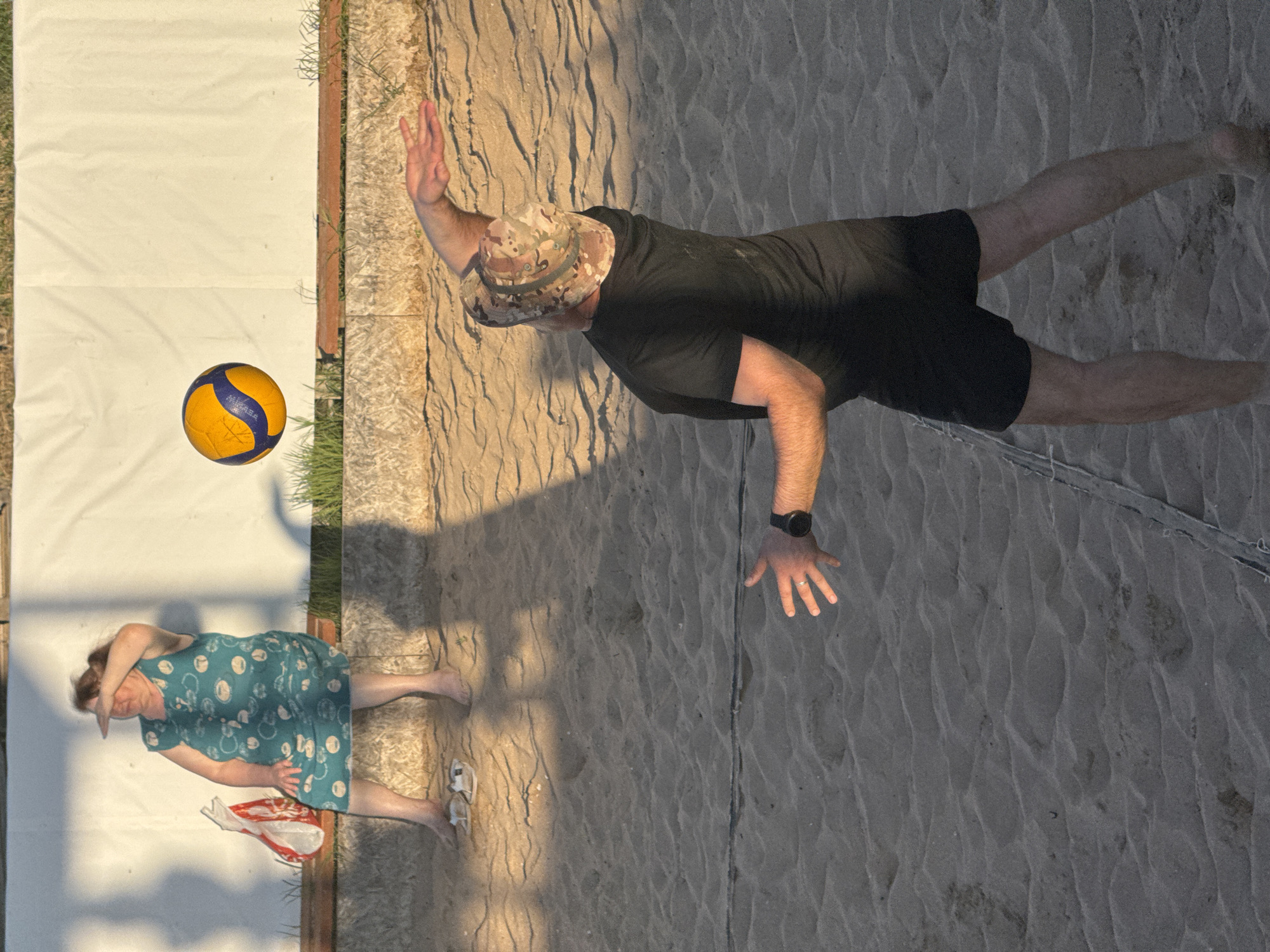 An outdoor daytime shot depicts a sandy volleyball court. In the background, a woman in a teal dress with a pattern of white circles sits on a bench, shielding her eyes from the sun. To her left is a red and white striped bag. In the foreground, a man wearing a black t-shirt, black shorts, and a camouflage hat is captured mid-action, with his right arm extended and his left hand raised. A yellow and blue volleyball is suspended in the air above him. The sand is textured with ripples and shadows cast by unseen objects. The scene appears to be located in the city of Shchelkino urban settlement, Ukraine.
