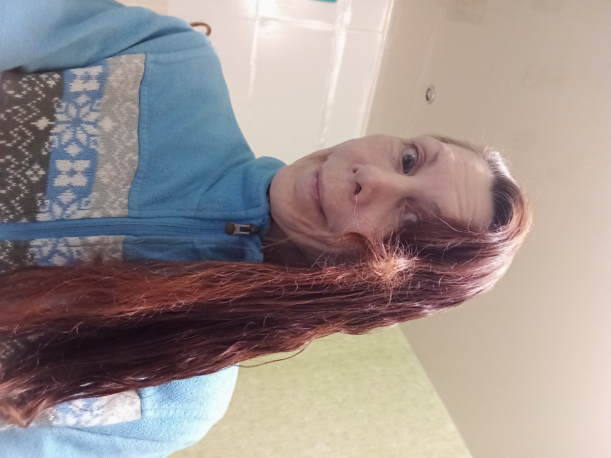 The media file shows a close-up, rotated portrait of a woman in what appears to be a bathroom or shower area. She is wearing a blue fleece jacket with a patterned insert on the chest featuring a snowflake motif in white, blue, and gray. Her hair is long, reddish-brown, and appears wet, with some strands sticking out. The background is a light-colored wall, possibly tiled, and a lighter colored wall with a subtle pattern or texture that could be a shower curtain or wall panel.
The woman's face is partially visible, angled towards the camera. She has fair skin, and her expression is somewhat neutral, with her eyes open and looking forward. The lighting suggests an indoor environment, possibly with artificial light.
The scene lacks any strong indicators of a specific time of day or weather. However, the wet hair and the background elements suggest the activity taking place is likely related to personal hygiene, such as showering or washing hair. The setting appears to be a domestic bathroom. There is no visible text in the image. The location context provided is Blagoveshchensk, Russia, but this cannot be visually confirmed from the image itself.