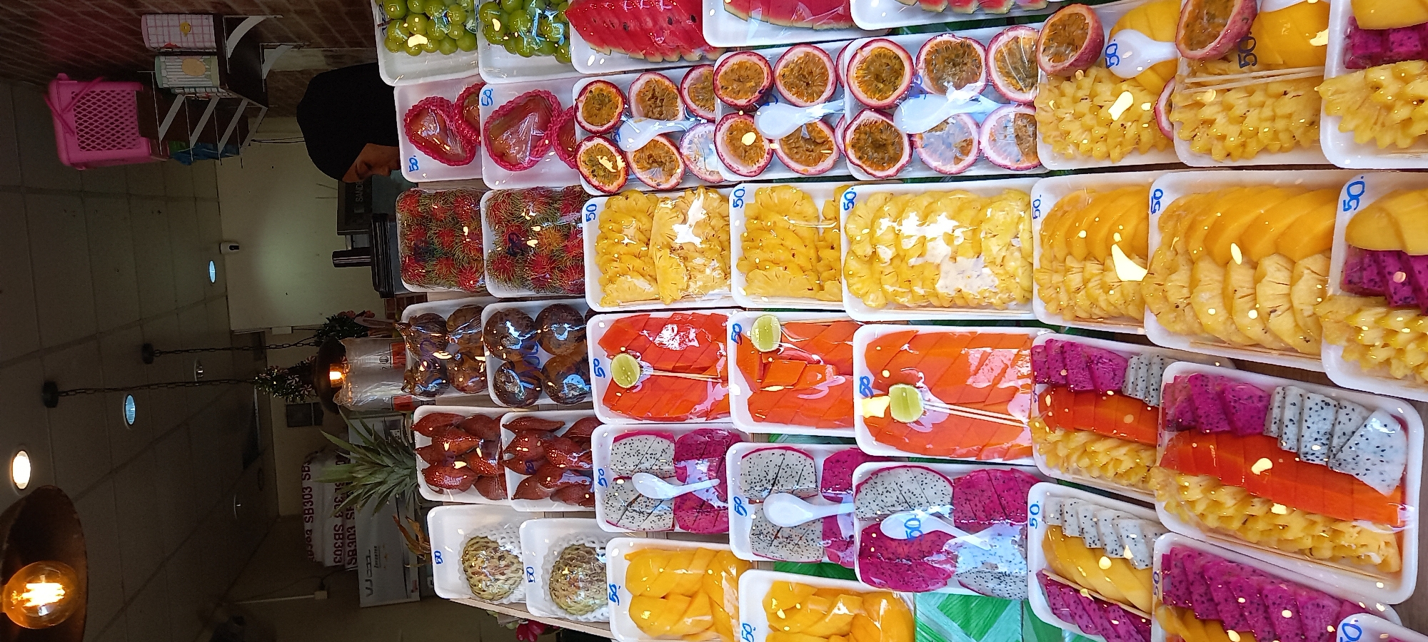 The image shows a display of prepared fresh fruit in the city of Ban Ao Nang, Thailand. The fruit is packaged in white plastic trays and covered in plastic wrap. The fruits include watermelon, passionfruit, pineapple, mango, papaya, dragon fruit, rambutan, longan, lychee and custard apple. Each tray has a sticker on it that appears to be pricing information. The fruit appears to be being sold in a market or store with various fixtures and equipment in the background. A person is standing in the background, wearing a black head covering.