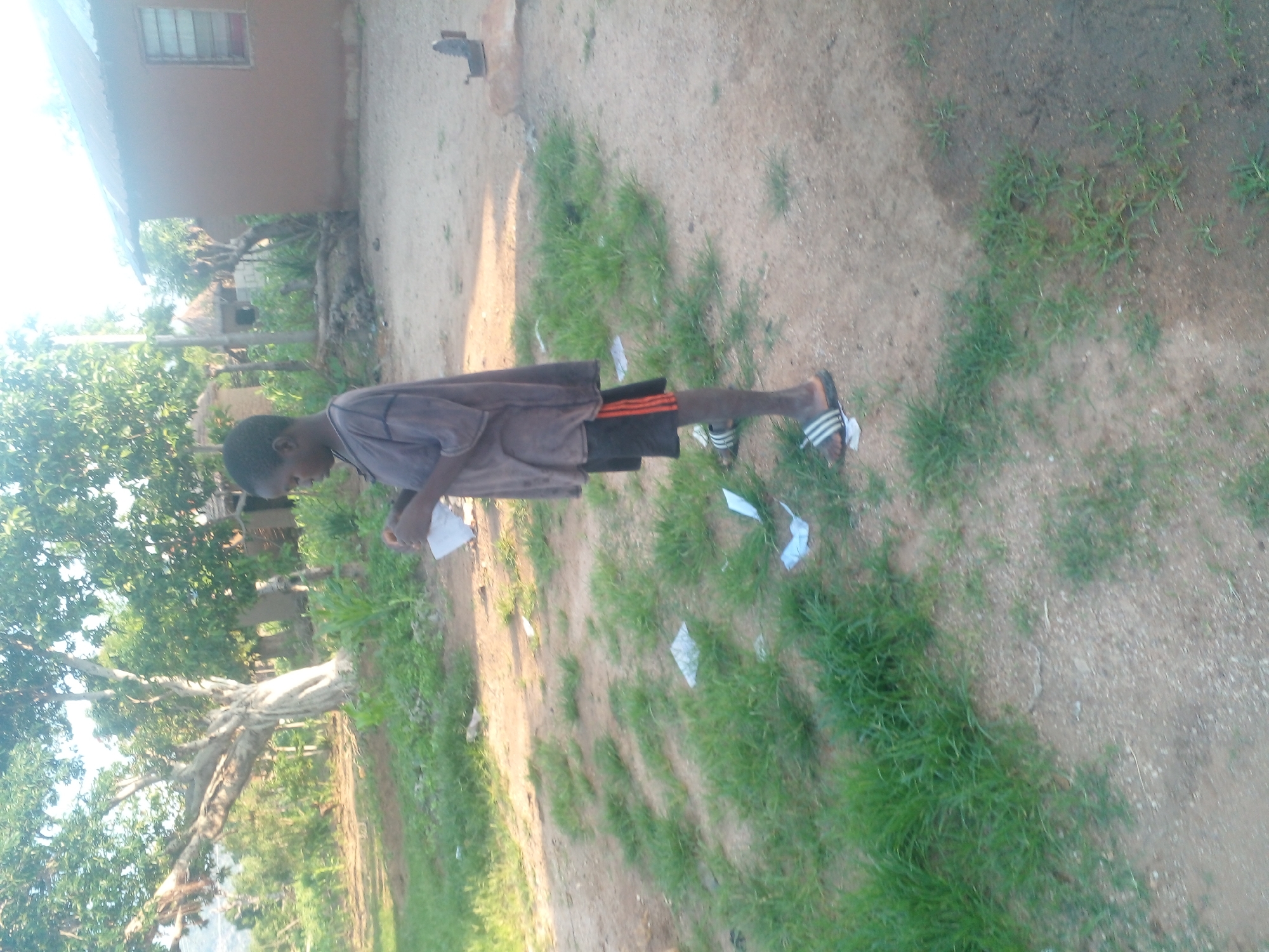 A young boy stands on a dirt path in what appears to be a rural village setting. He is wearing a dark gray t-shirt and black shorts with orange trim, and flip-flops. The boy is holding a white piece of paper in his hands. To his left, there is a large tree with bare branches and green foliage. In the background, there are several huts with thatched roofs and a brown building with a metal roof and a window. The ground is covered with dirt, patches of grass, and some scattered white paper. A rusty iron is visible on a concrete block in the background. The scene is lit by natural sunlight, casting shadows on the ground.