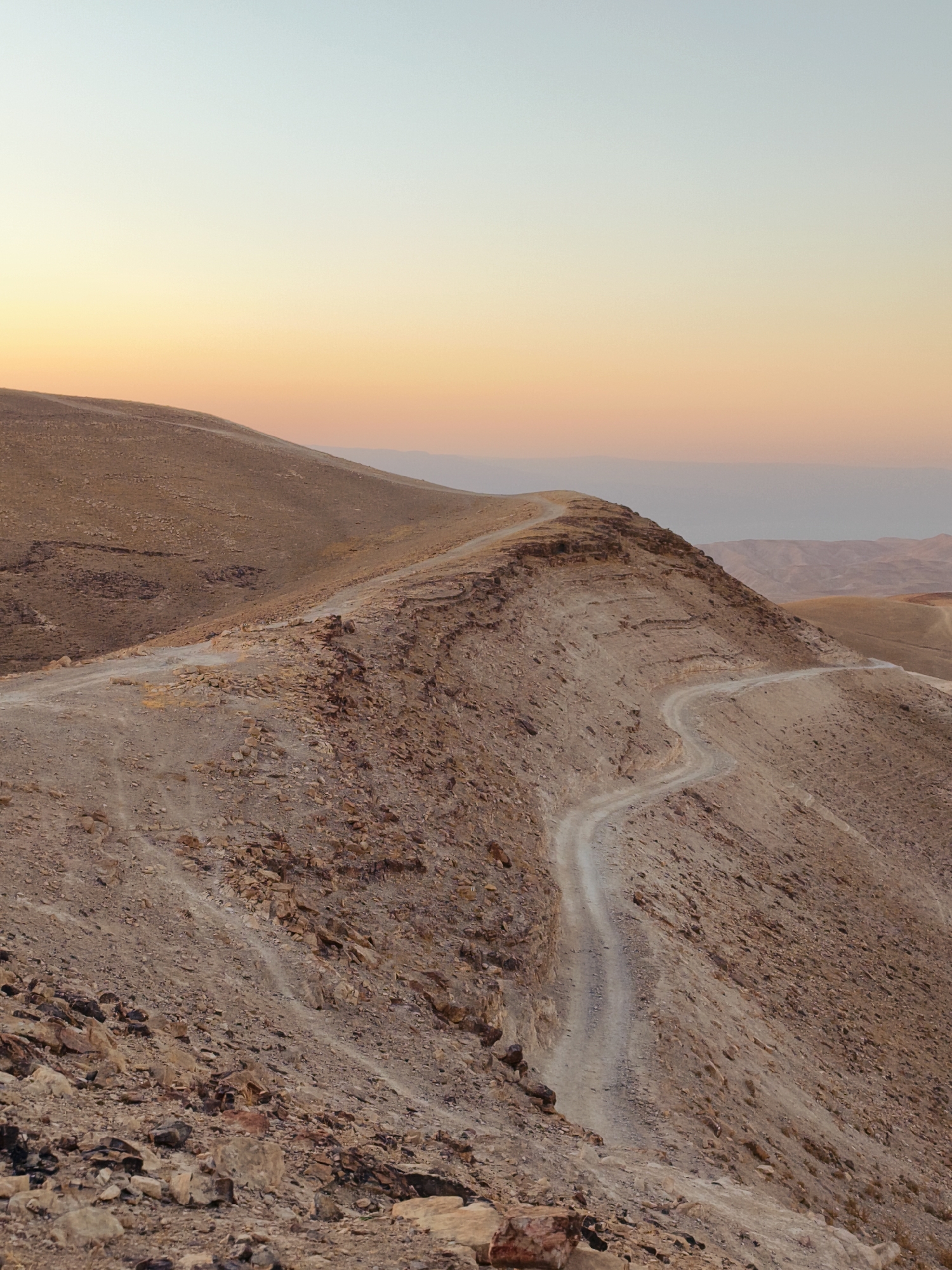A dirt road winds its way up a barren, rocky hillside in a desert landscape. The sky above transitions from a pale blue at the top to a warm orange and yellow gradient near the horizon, suggesting either sunrise or sunset. The terrain is characterized by dry earth, scattered stones, and sparse vegetation. In the distance, additional rolling hills and mountains are visible, fading into a hazy atmosphere. The road appears to be a single-lane track, carved into the side of the hill. The scene is devoid of any people or structures. The overall impression is one of natural, rugged beauty and solitude. The location is the city of Keidar, Palestinian Territories.