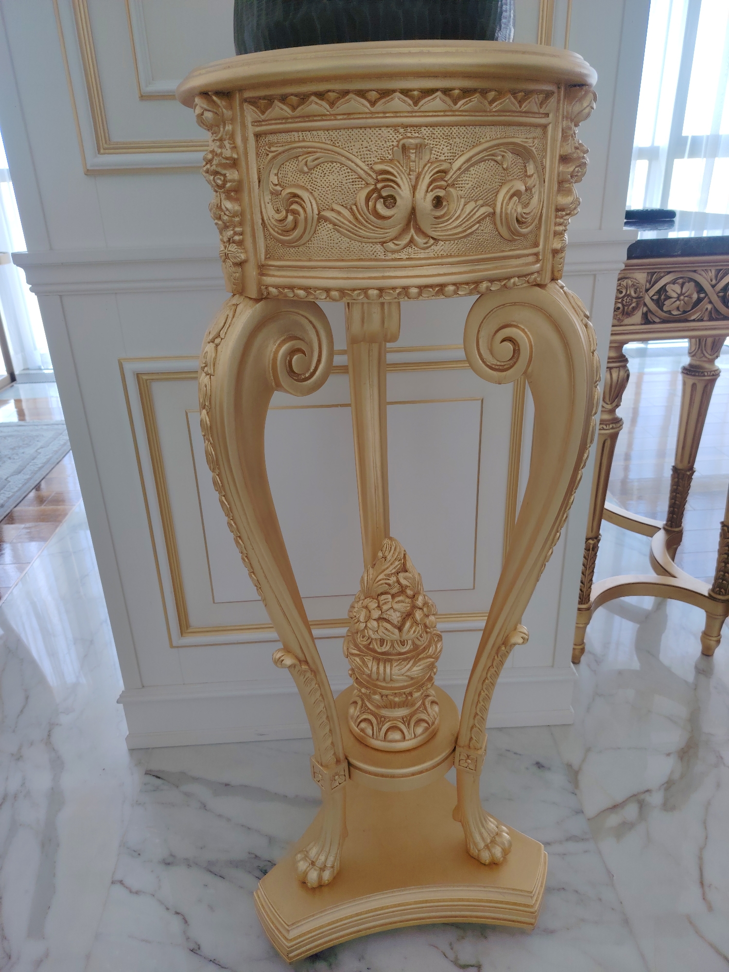 The image displays a close-up of an ornate, gold-colored pedestal table, likely a decorative accent piece. The table features intricate carvings and scrollwork on its top and legs, with a detailed floral motif at the base of the central support. It is positioned on a polished marble floor, reflecting the light. In the background, a white wall with gold trim and molding suggests an indoor, likely residential or formal setting. To the right, another similar gold table with a dark top is partially visible. The lighting is bright, possibly from natural light filtering through a window, indicated by the sheer curtains visible in the far background. There are no people or animals present in the scene. The overall impression is one of opulence and classic design. This scene is not located in Konigil, Uzbekistan.