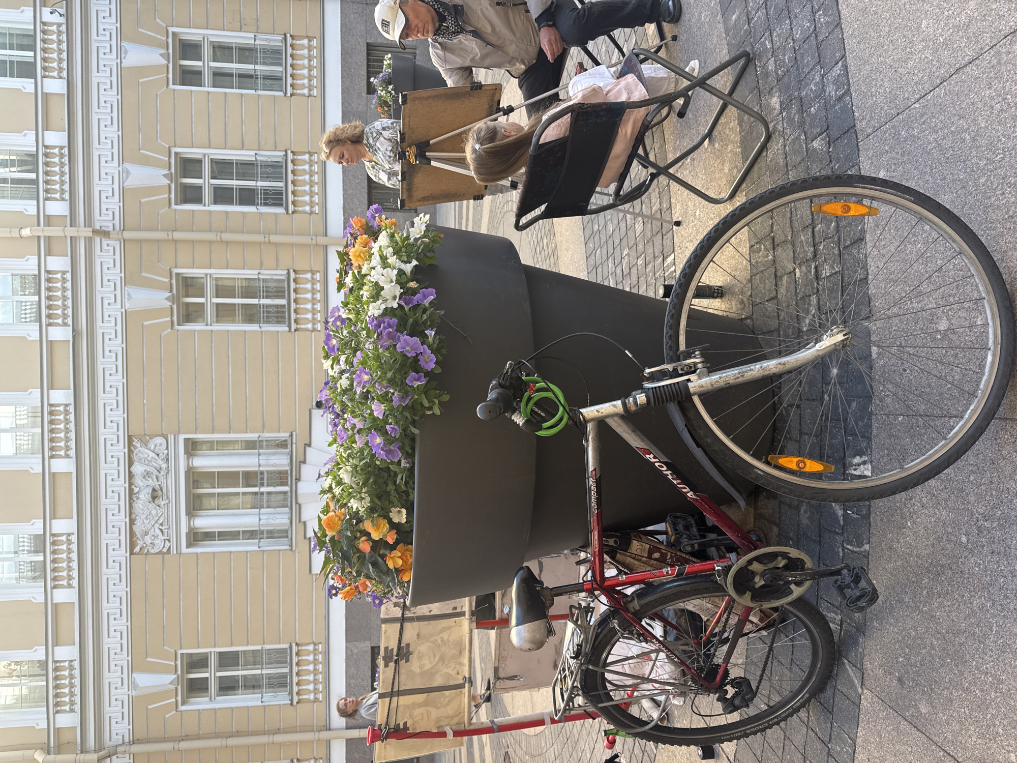In Saint Petersburg, Russia, on a cobblestone street lined with buildings featuring ornate facades and balconies, an outdoor scene unfolds. A large, dark planter overflows with vibrant orange, yellow, and purple flowers, dominating the foreground. To the left, a red and silver bicycle leans against the planter, its front wheel prominently displayed. In the mid-ground, a man, wearing a cap and a scarf, is seated on a folding chair, intently painting a young girl who sits facing him, also on a folding chair. The girl is dressed in a light pink outfit. Behind them, a woman with blonde, curly hair stands, observing the artistic activity. Another easel and a portable art studio setup are visible further back, suggesting an artistic gathering or a public art demonstration. The architectural style of the surrounding buildings indicates a historic district within the city.