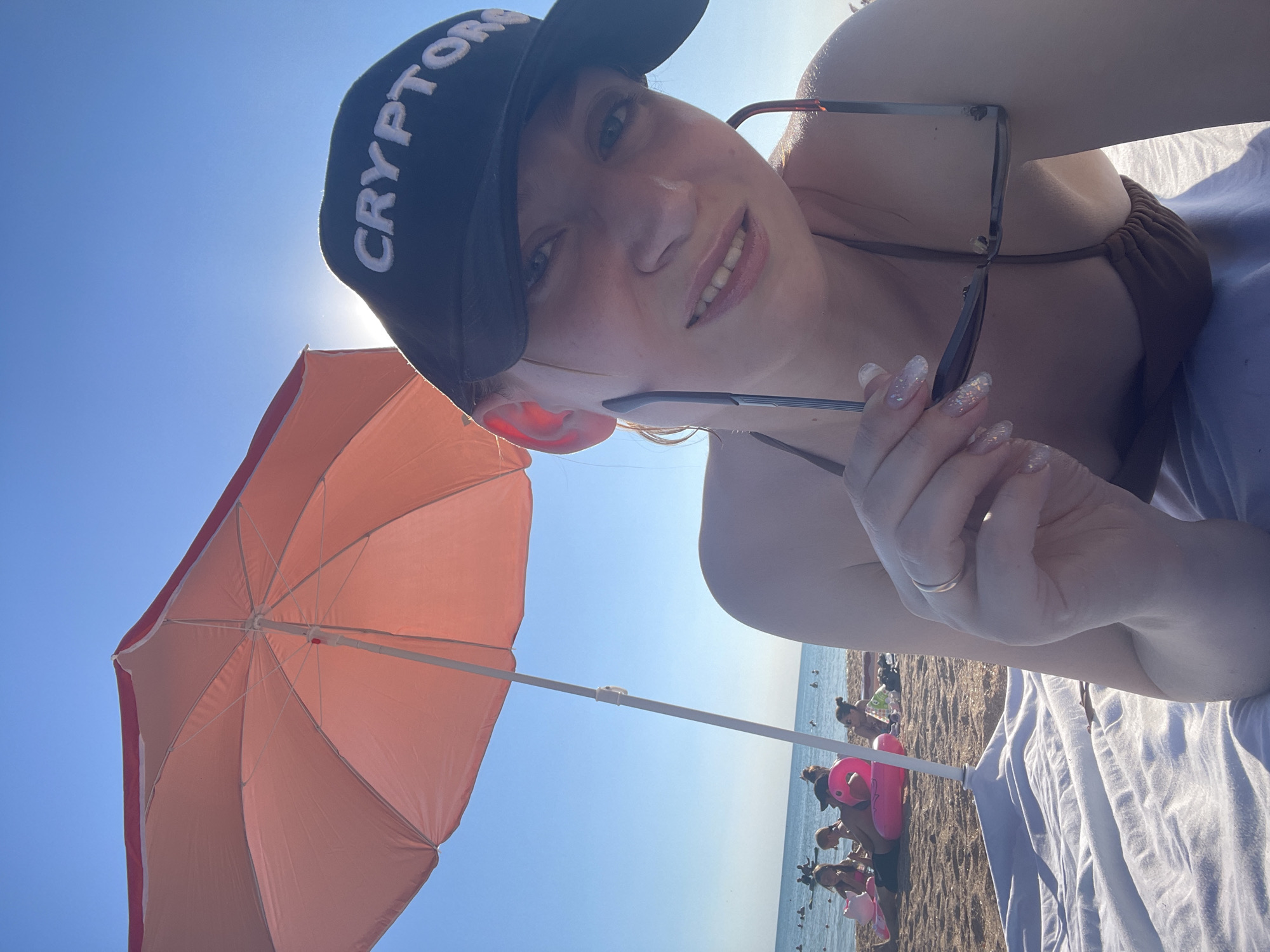 A woman lies on a beach under a pink umbrella, looking at the camera and holding a pair of glasses. She is wearing a dark bikini top and a black baseball cap that says "CRYPTOR" in white letters. The beach is sandy, and the ocean is visible in the background. Other people are sitting and swimming in the water. The sky is clear and blue, and the sun is shining. The location is Blagoveshchenskaya, Russia.