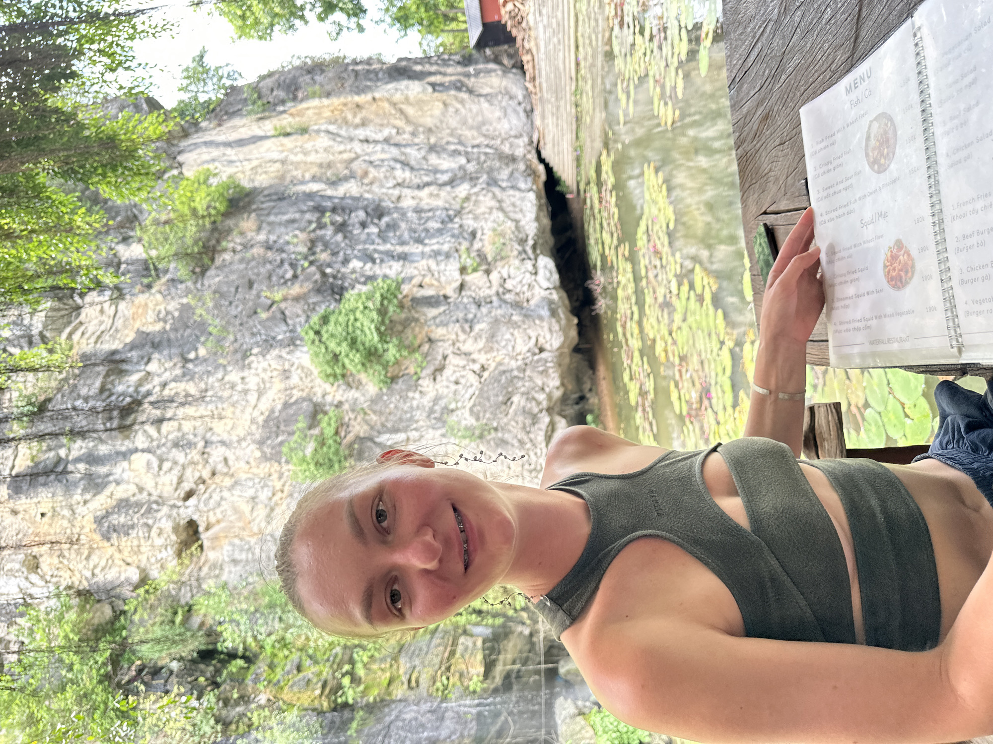 In Nam Hoa Lư Ward, Vietnam, a young woman with blonde hair pulled back and braces smiles at the camera. She wears a green, cut-out crop top and dark pants. She is seated at a wooden table outdoors, looking at a menu. The table is positioned in front of a pond covered with lily pads. Behind the pond, a large, rocky cliff face rises, covered in patches of green vegetation and vines. To the far right of the cliff, a small red building with a dark roof is visible. The overall scene suggests a natural, scenic dining environment.
