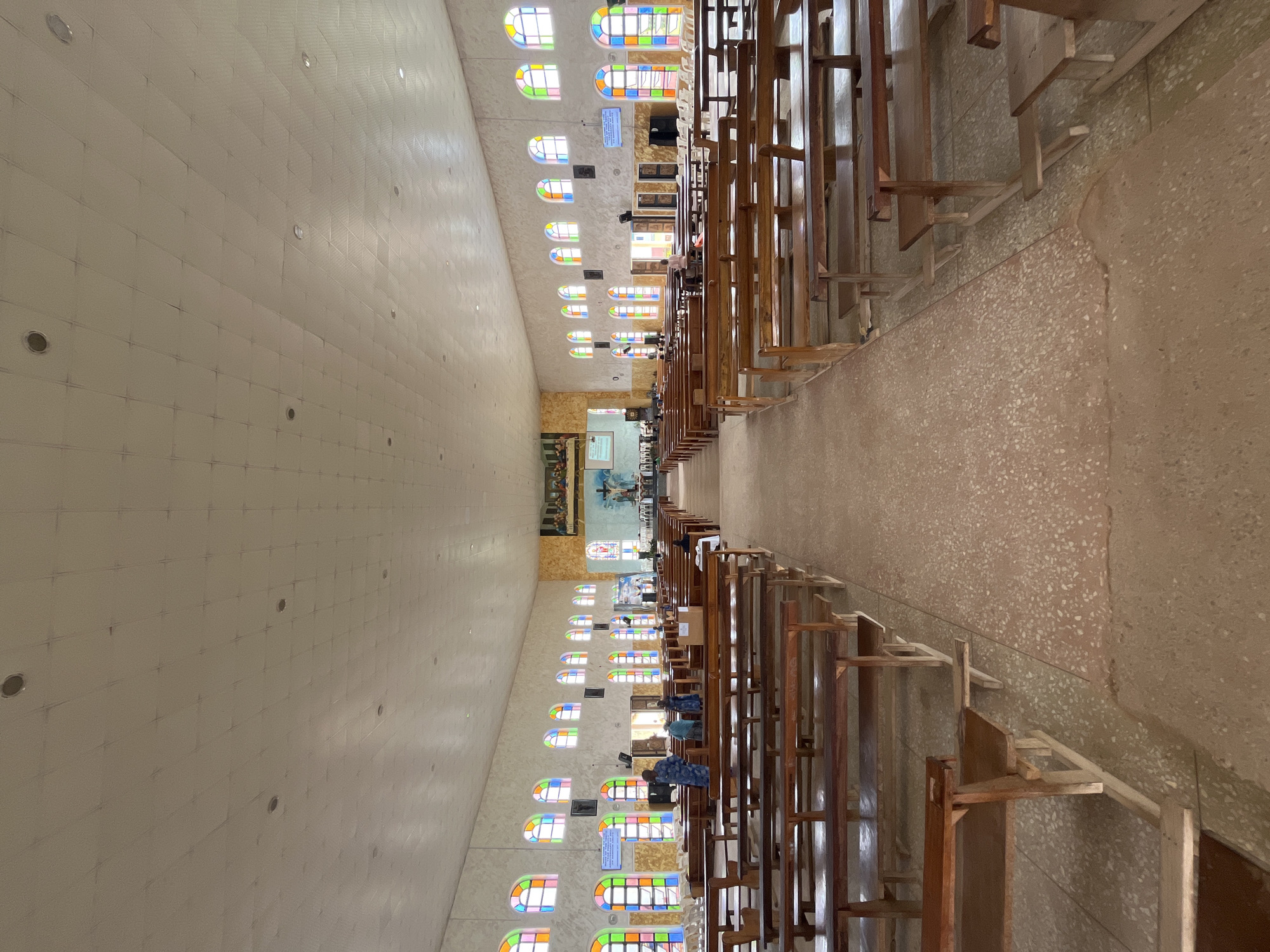 The image shows the interior of a large, brightly lit church in Vom, Nigeria. The church features rows of wooden pews on either side of a central aisle. The walls are lined with arched stained-glass windows in various colors, including red, yellow, green, and blue. At the far end of the church, a large mural depicting a religious scene is visible above the altar. Several people are present in the church, including individuals standing near the front and seated in the pews. The ceiling is white and appears to be made of tiled panels, with several circular light fixtures embedded in it. The floor is a light brown, speckled material. The overall atmosphere is one of reverence and quiet contemplation.