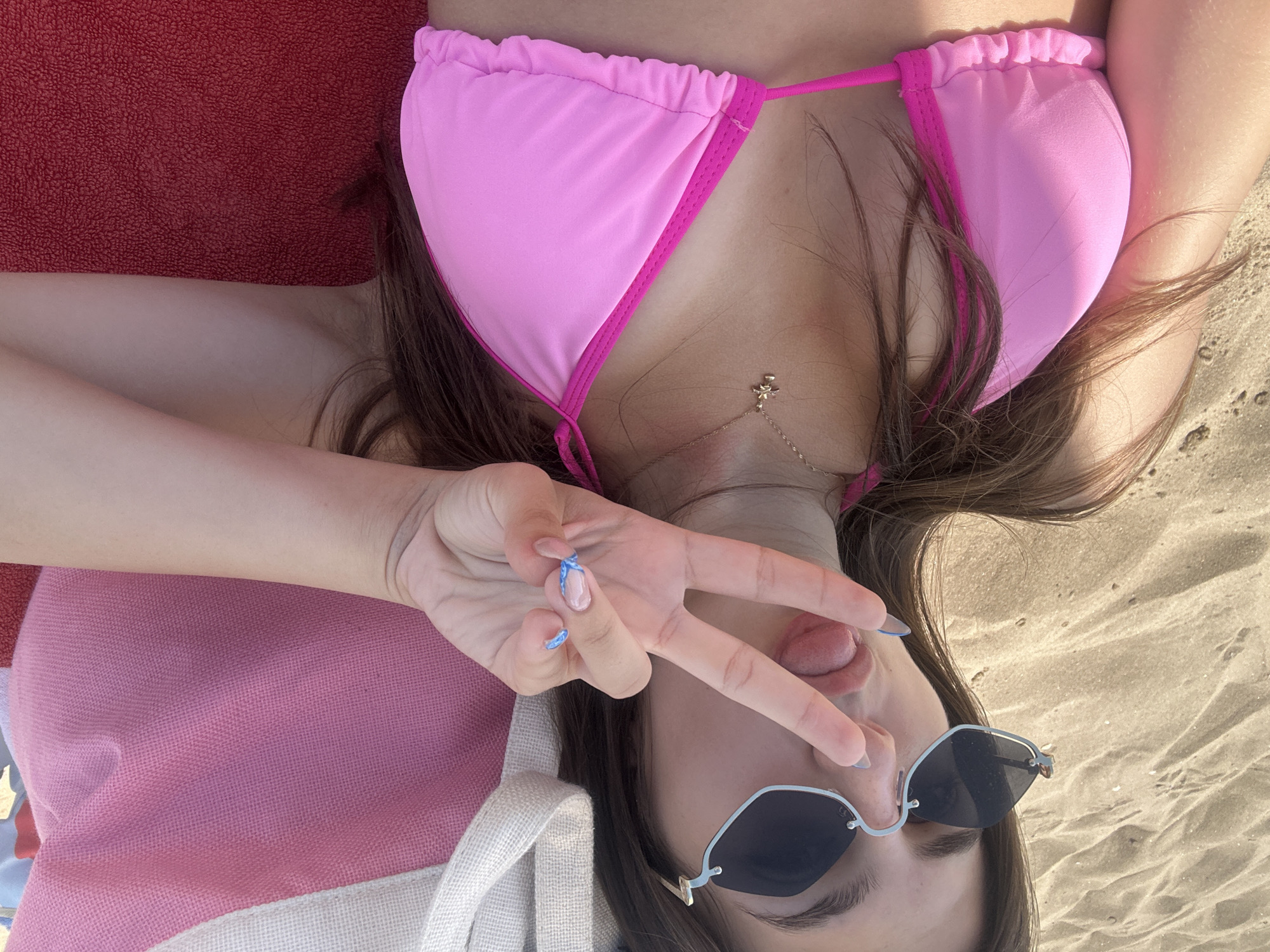 A young woman with long brown hair and sunglasses is lying on a beach in Nesebar, Bulgaria. She is wearing a pink bikini top and has her tongue sticking out while making a peace sign with her fingers. Behind her, a pink and white beach bag is visible. The sand is visible in the foreground and background.