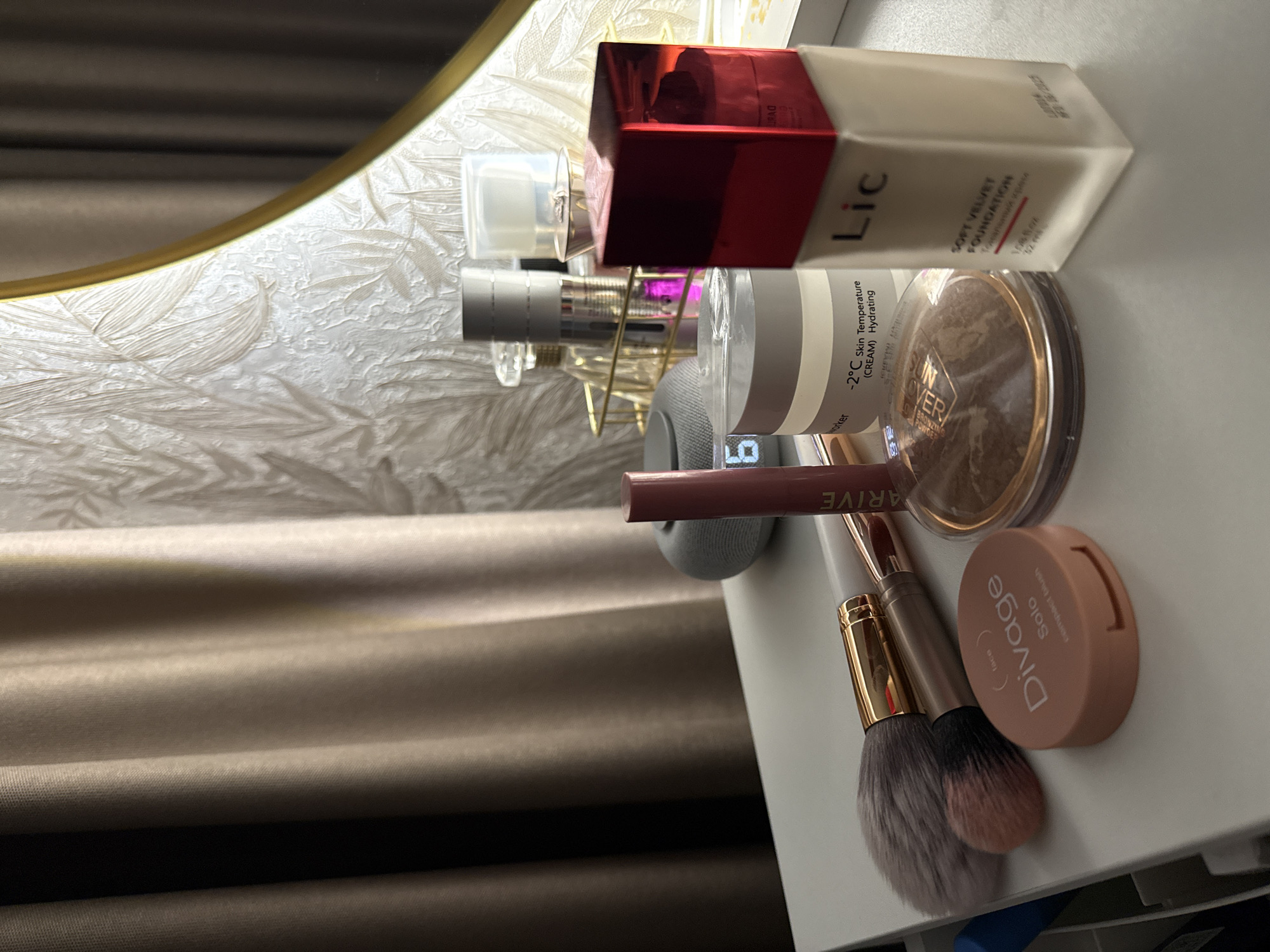 The image shows a collection of cosmetics on a white surface. Products include a "Lic Soft Velvet Foundation," a bronzing powder labeled "Sun Lover," a Divage Solo compact blush, and what appears to be a Thrive lip product. Makeup brushes are also visible. The scene includes a mirror with a decorative frame and beige curtains in the background. It is not possible to determine the location in Izhevsk, Russia, from the provided image.