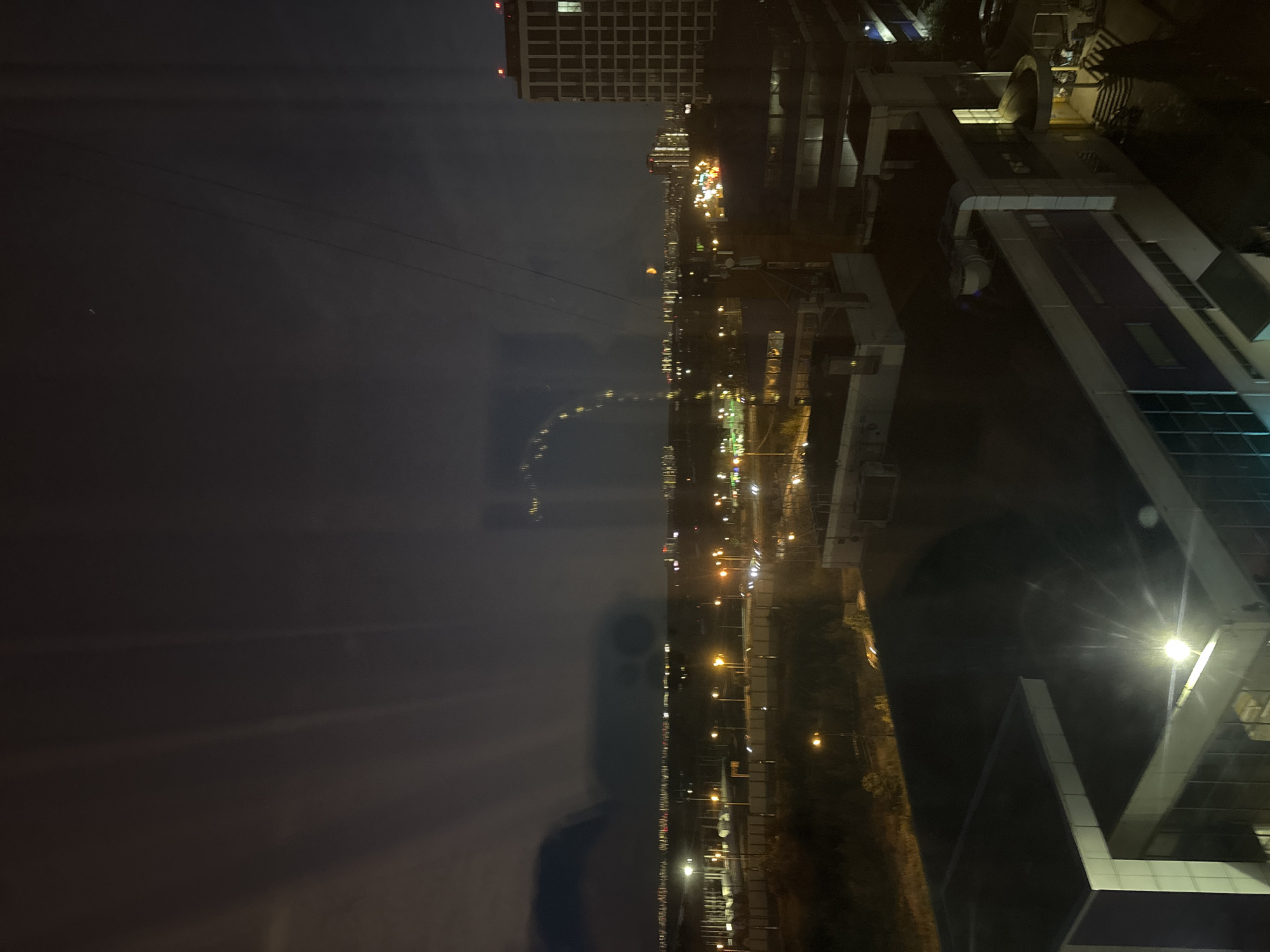 This is a nighttime aerial view of an industrial or commercial area in Odintsovo, Russia. The image captures several buildings with illuminated windows and external lighting, suggesting ongoing activity or security measures. In the foreground, two low-rise buildings with flat roofs are visible, one with a prominent white structure and bright exterior lighting. Further in the background, a taller, multi-story building stands out, its windows lit. The landscape is dotted with streetlights and other sources of illumination, creating a scattered glow across the dark terrain. Reflections on the surface from which the image is taken are visible, particularly in the upper left and central portions of the frame, distorting some of the distant lights and creating amorphous shapes. A thin wire or cable is visible stretching across the sky. The overall atmosphere is one of a sprawling urban or suburban development at night, with the presence of man-made structures dominating the scene.