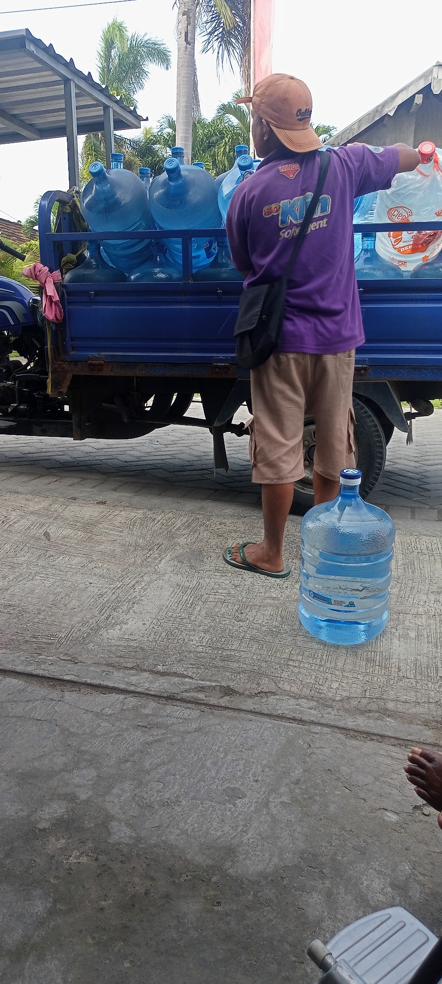 In Gempolklutuk, Indonesia, a person wearing a purple shirt with the "Sok Ken Soft Gent" logo and a brown baseball cap stands next to a blue truck laden with large blue water containers. The truck also carries a few transparent water bottles. The individual is wearing beige cargo shorts and flip-flops. A large, partially filled water jug rests on the ground in front of the truck. The scene appears to be outdoors, possibly a roadside or delivery area, with a concrete surface and some vegetation visible in the background.