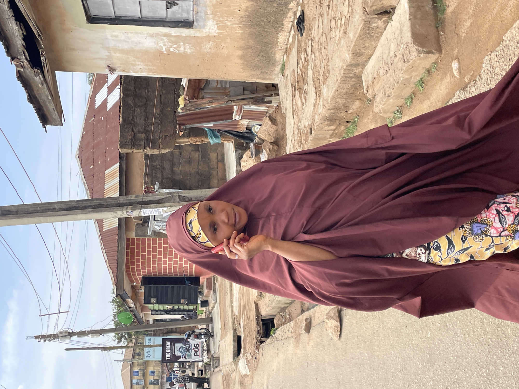 The image, presented rotated on its side, captures a young woman standing on a street in what appears to be an urban or semi-urban area, consistent with Jos, Nigeria. She is positioned in the foreground, looking directly at the camera with a neutral to slightly smiling expression. She wears a maroon head covering (hijab) and a flowing maroon outer garment, underneath which a vibrant yellow dress with a colorful floral pattern is visible. In her right hand, she holds a red object, likely a mobile phone, to her ear or near her mouth.
The background showcases a bustling street scene under a clear, bright sky. To the left, utility poles with numerous electrical wires crisscross the frame. Several buildings line the street; some exhibit older, weathered concrete construction with visible brickwork and corrugated iron roofs, suggesting varying states of repair. The road appears paved, with a rougher, possibly unpaved section or drain running alongside where the woman stands. In the mid-ground on the left, a sign is visible with text "THE GREAT" and "POS," with "Cyber..." and "BUSINESS" also partially discernible. A person and a small motor vehicle can be seen in the distance near the sign. The photograph captures a candid moment of daily life under bright daylight conditions.