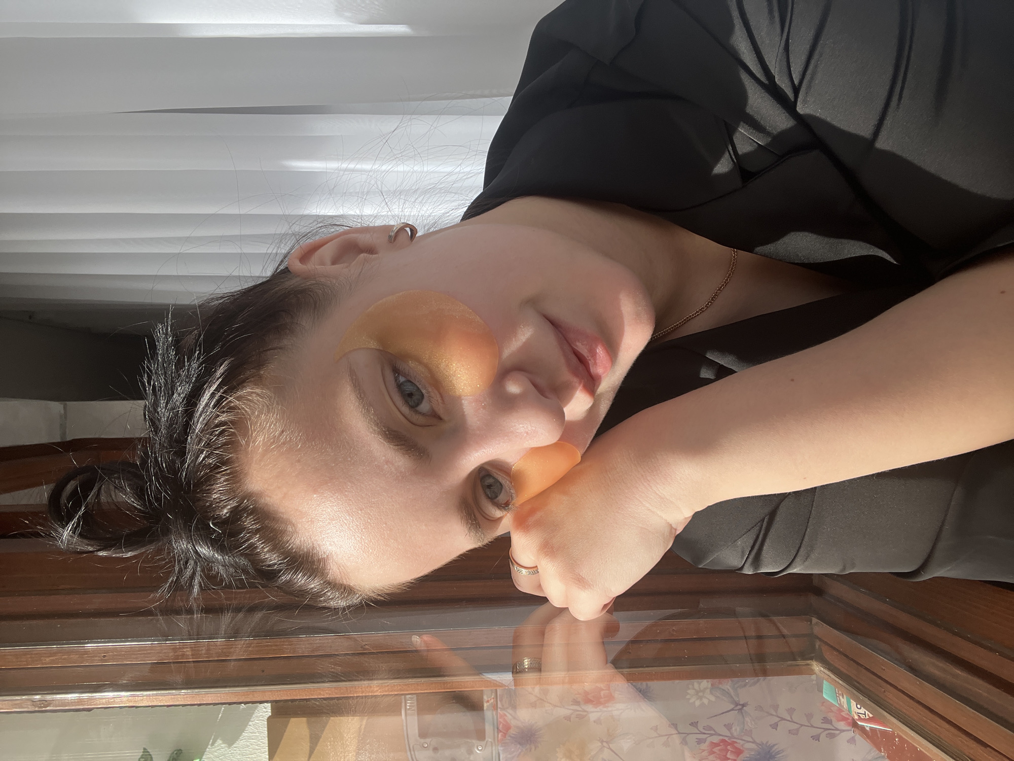 A woman with light skin and dark hair is lying on a glass surface with her head tilted to the left. She is wearing a black top and gold under-eye patches. Her left arm is bent and her fist is near her face. A gold ring is visible on her left ring finger. The glass surface reflects the woman's face and hand, as well as a floral patterned surface underneath the glass. Natural light is illuminating the scene, casting shadows on the woman and the background. There is no evidence to suggest the location is Марковский, Russia.