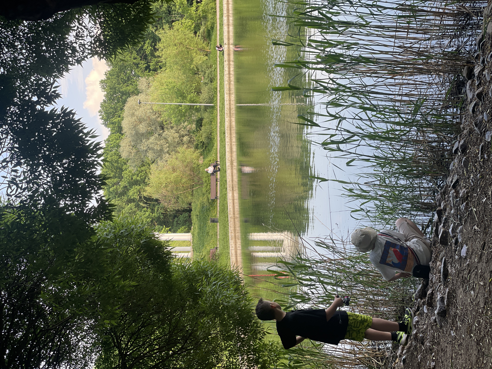 In Khimki, Russia, a boy in a black t-shirt and green shorts and a person in a white shirt and hat are near a lake. The boy is lying on the ground near tall grasses. The other person is crouching near the lake, possibly fishing. The lake's surface reflects the trees and sky. A paved walkway with benches runs along the opposite side of the lake. Trees surround the area, providing shade in the foreground and background.